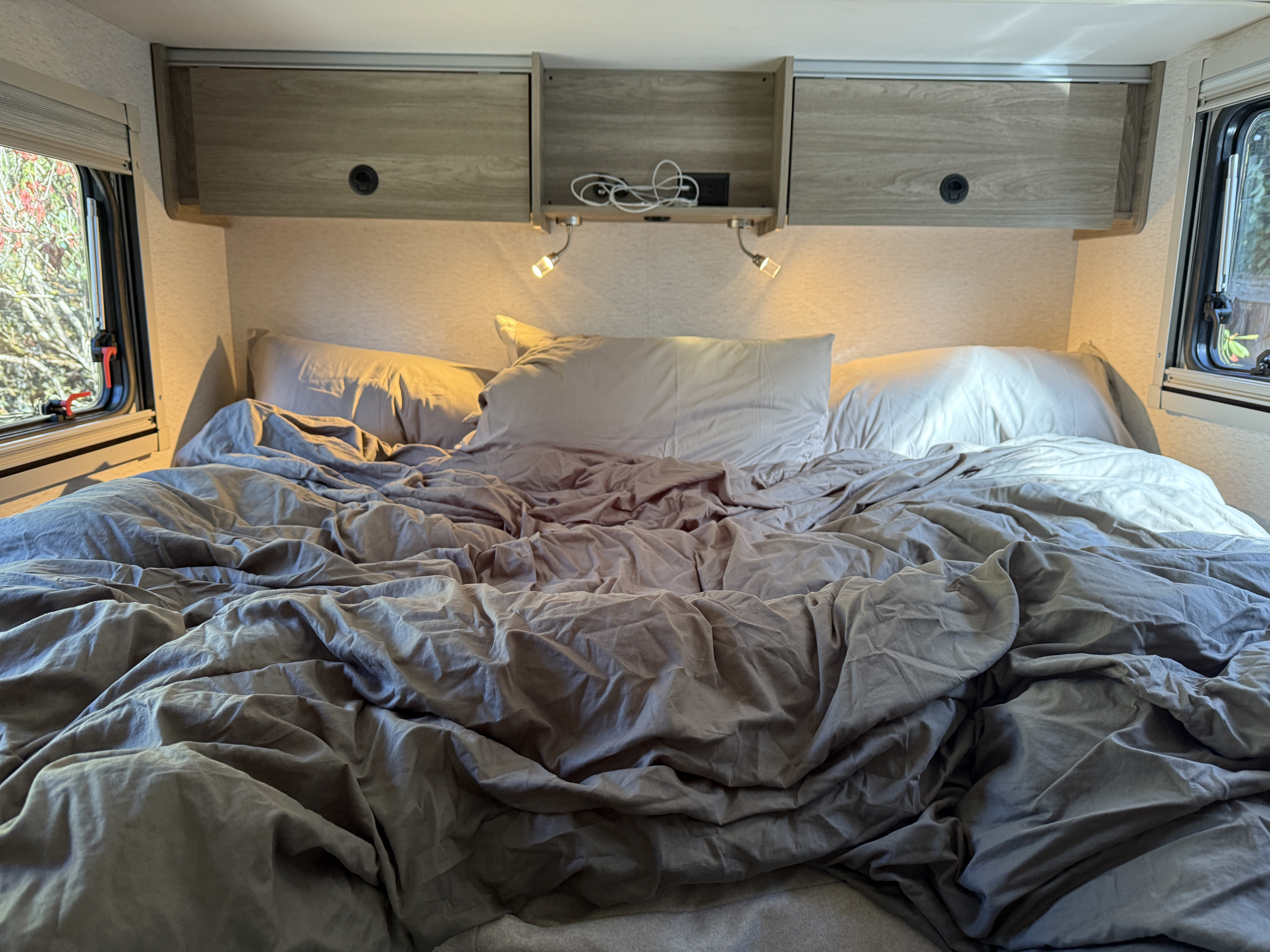 Unmade bed in a comfortable RV rental, featuring reading lights, overhead storage, and windows with outdoor views.. Winnebago Ekko 2025