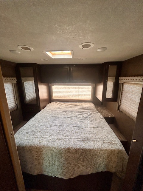 This RV rental bedroom features a patterned bed, dark wood cabinets above, and windows with blinds on each side.. Thor Four Winds 2020