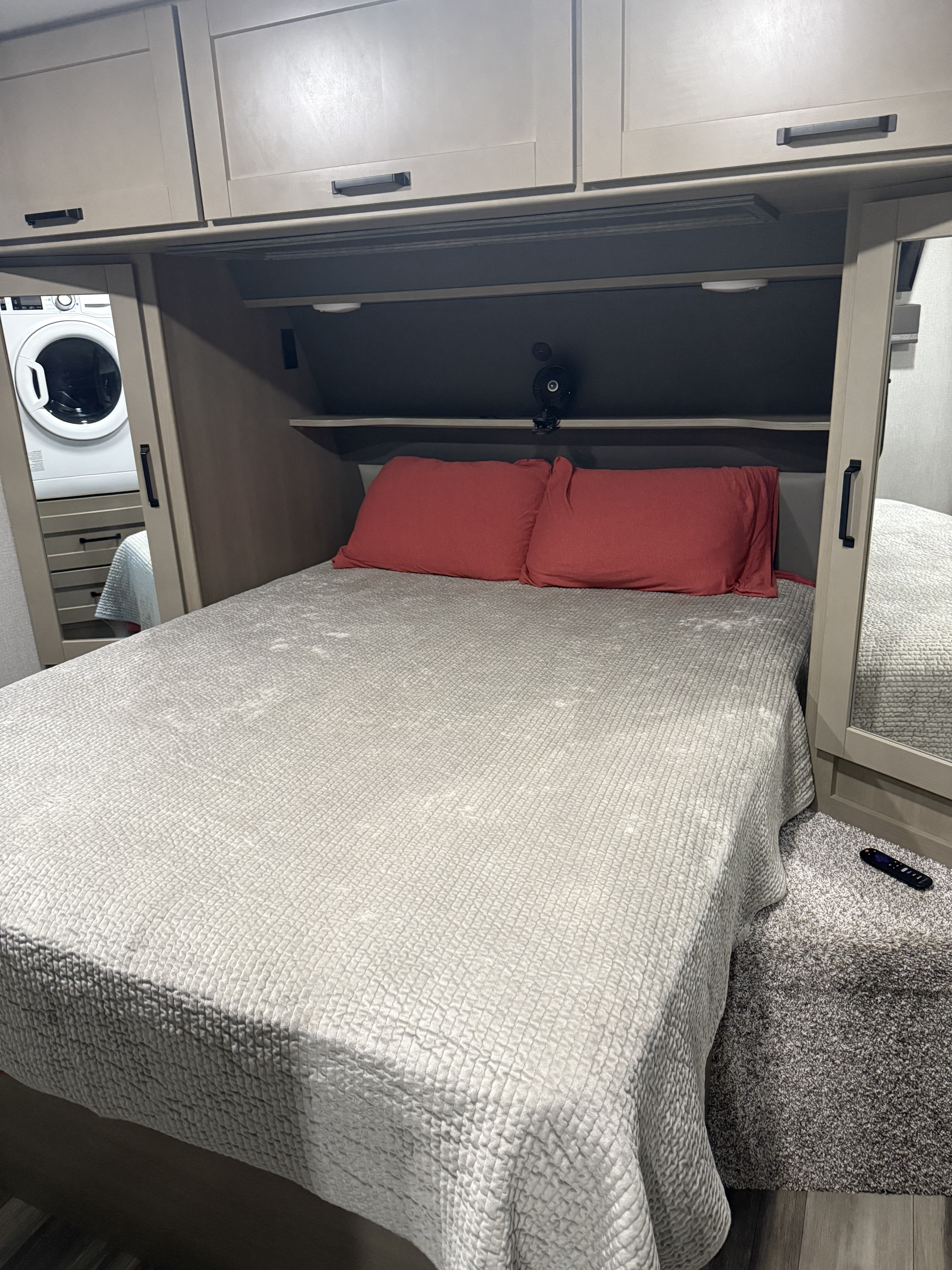 RV rental bedroom featuring a bed with red pillows, overhead storage, and a reflected washing machine. Compact travel space.. Grand Design Reflection 2024