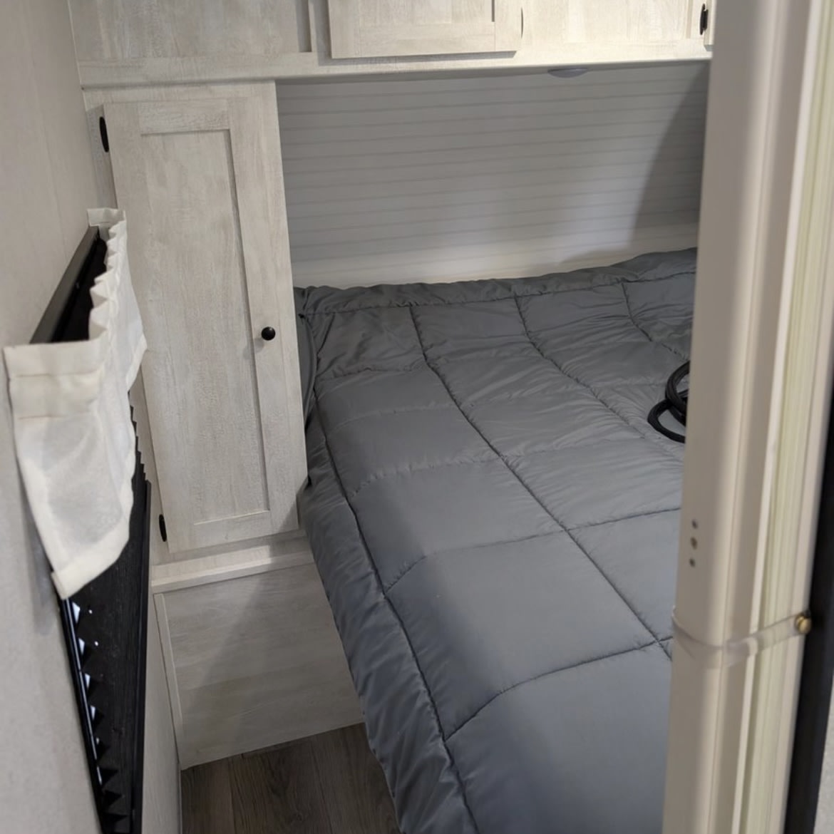 Cozy RV rental bedroom featuring a grey comforter, light wood cabinetry, and a small curtain.. Coachmen I-5 Edition 530QB 2024