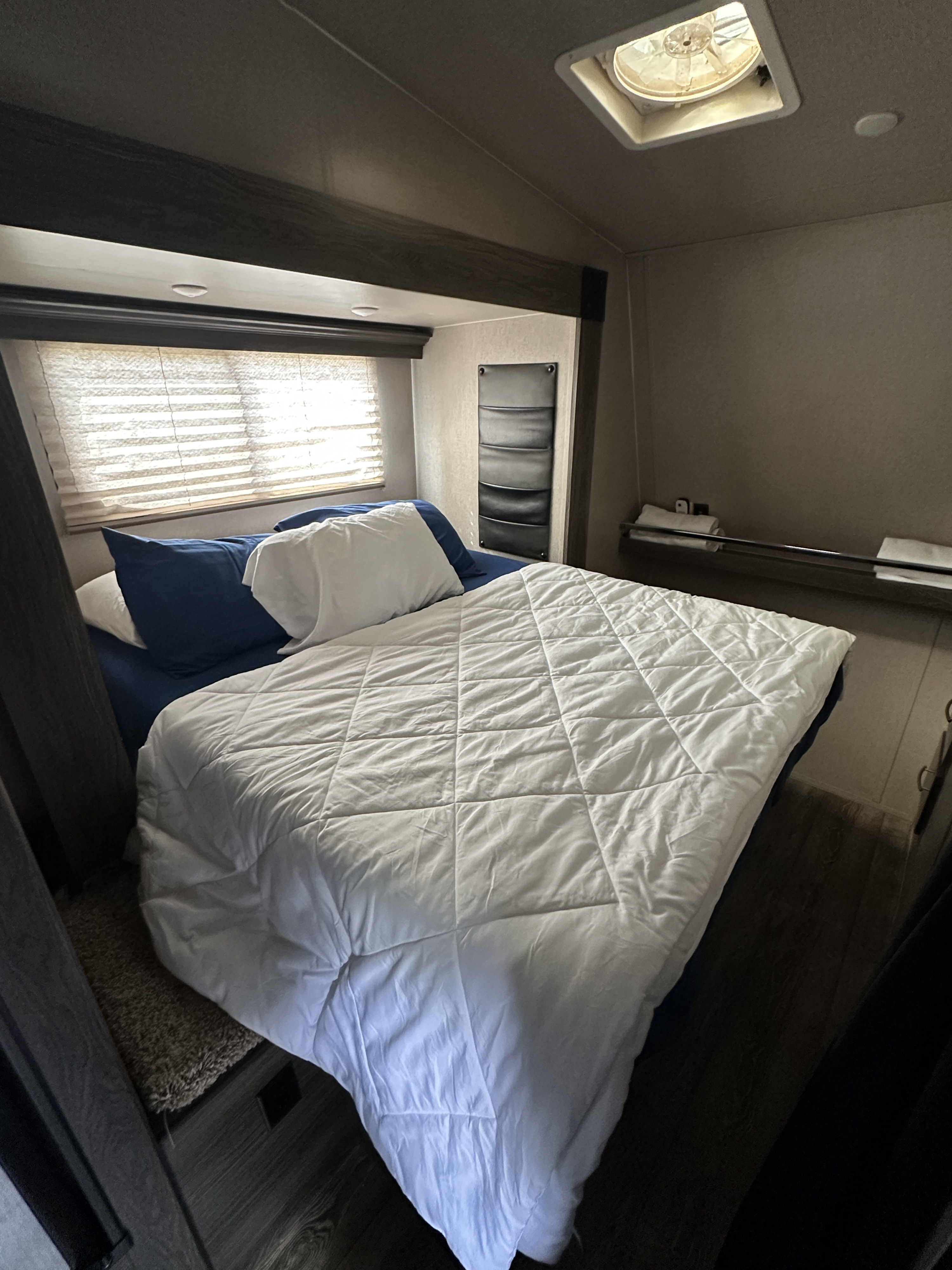 Cozy RV rental bedroom interior features a comfortable bed, window blinds, overhead vent, and shelf. Ideal for travel.. Genesis Supreme Rv Vortex 2019
