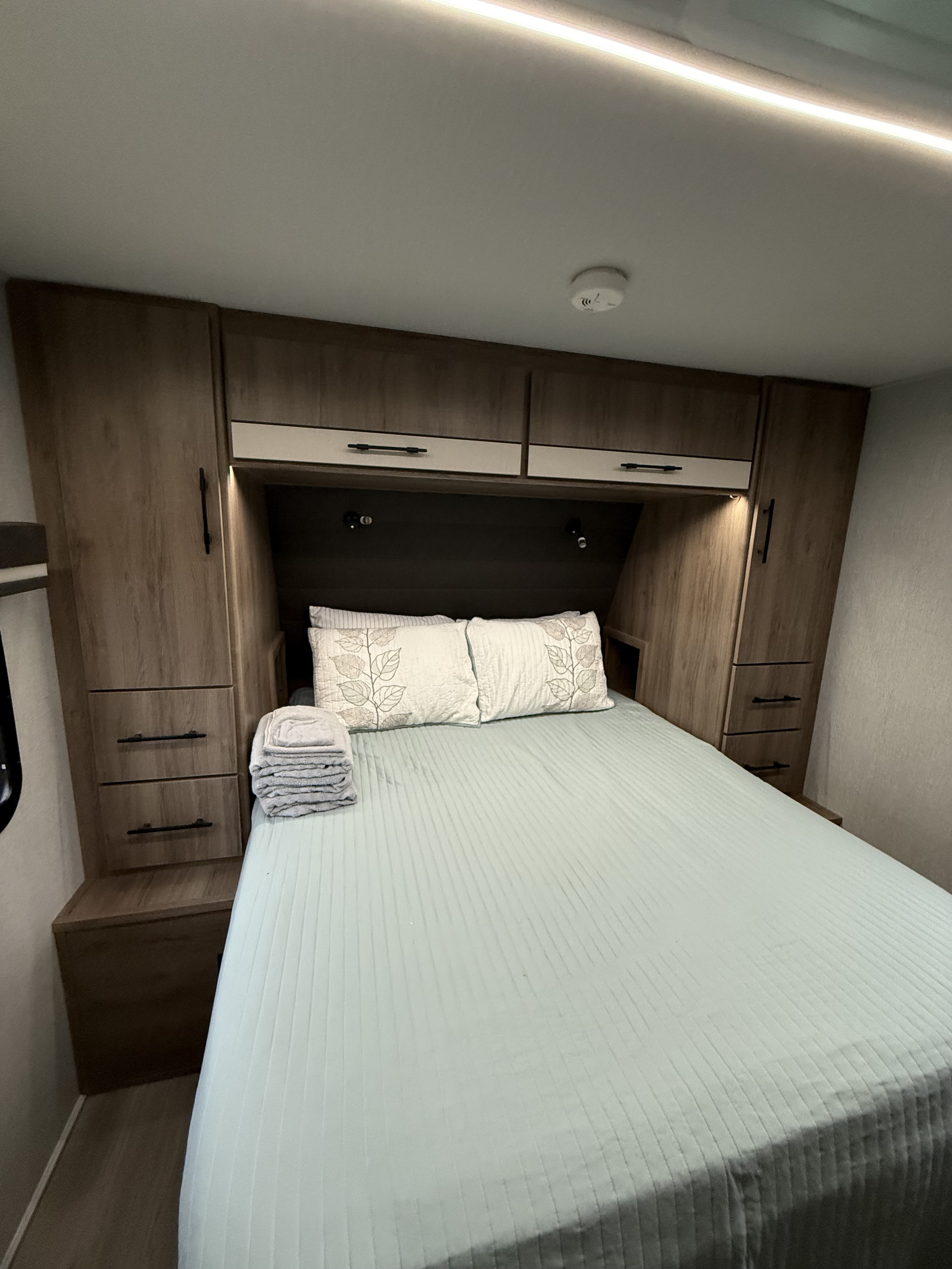 RV rental bedroom: A comfortable bed, light blue quilt, integrated wooden storage, and a ceiling smoke detector.. Grand Design Imagine 2025