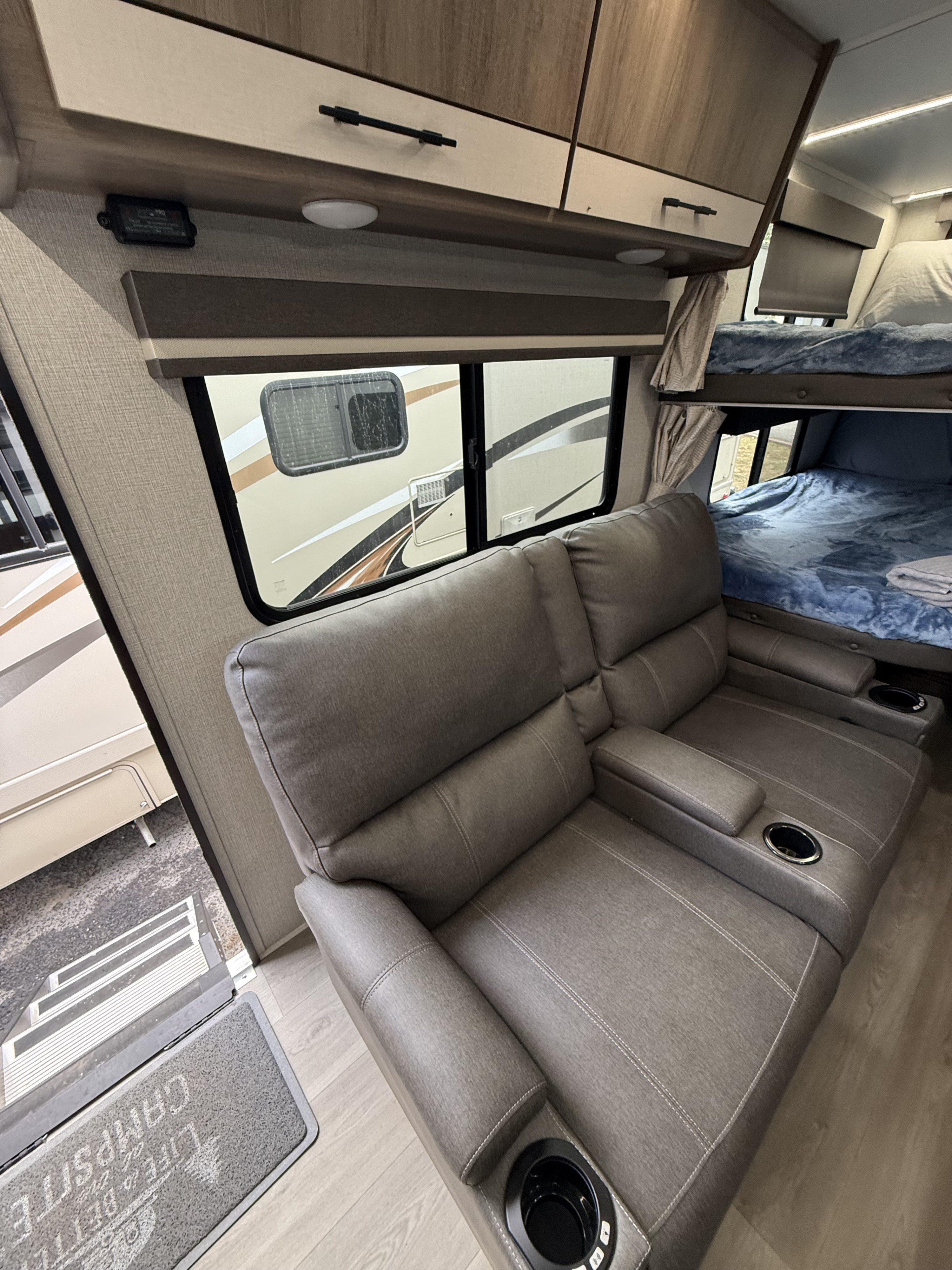 RV rental interior: a gray recliner sofa with cupholders, bunk beds, and windows showing another camper outside.. Grand Design Imagine 2025