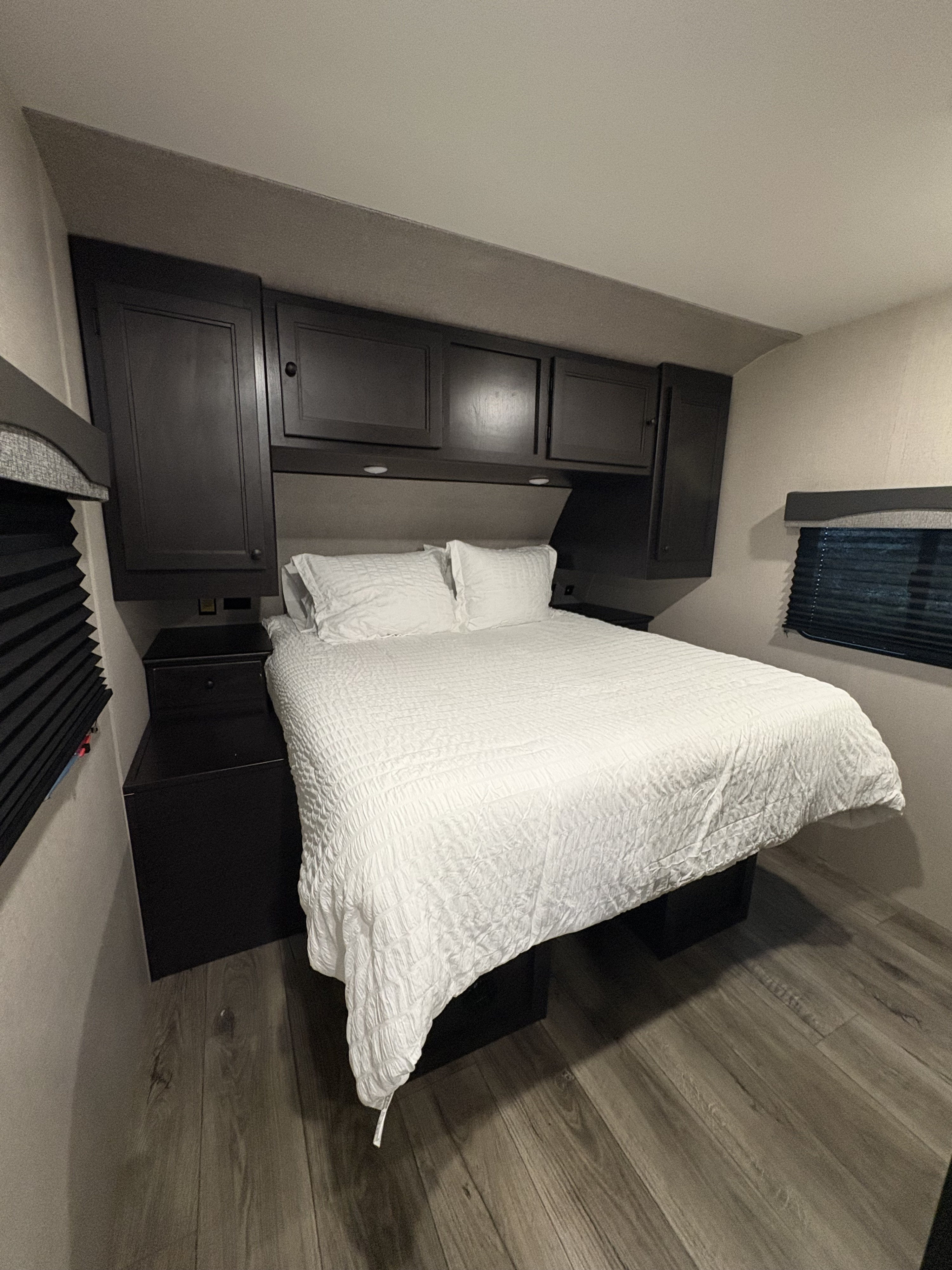 Inside an RV rental bedroom: a queen bed with white bedding, dark built-in cabinets, and side window blinds.. Highland Ridge RV Range Lite 2021