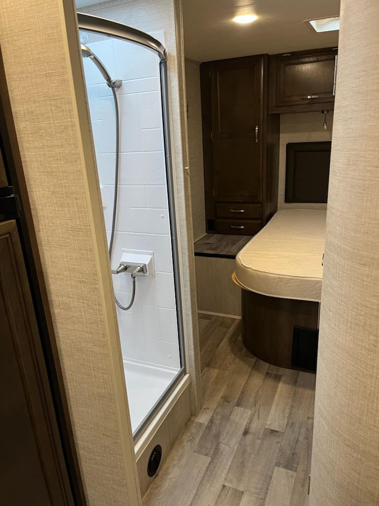 This RV rental interior features a compact shower stall on the left and a cozy bedroom area with dark wood cabinets.. Jayco Redhawk 29XK 2023