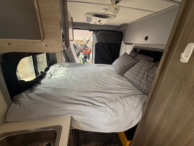 Interior view of an RV rental: a comfortable bed with grey bedding and pillows, and a small sink area.. Winnebago Solis 2022