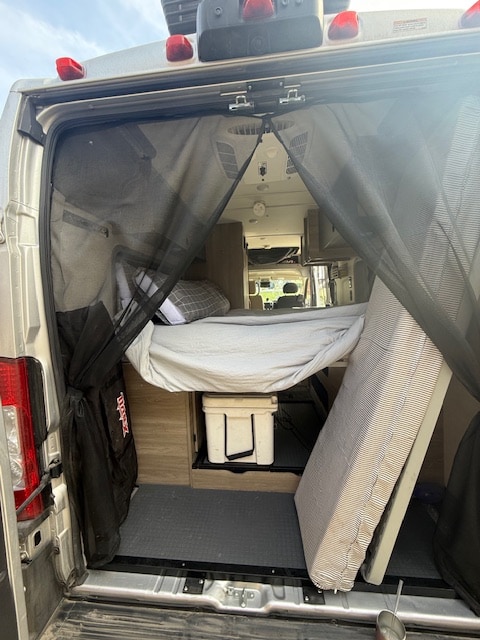 View into an RV rental from the open rear, revealing a mesh screen, raised bed, and interior storage including a cooler.. Winnebago Solis 2022