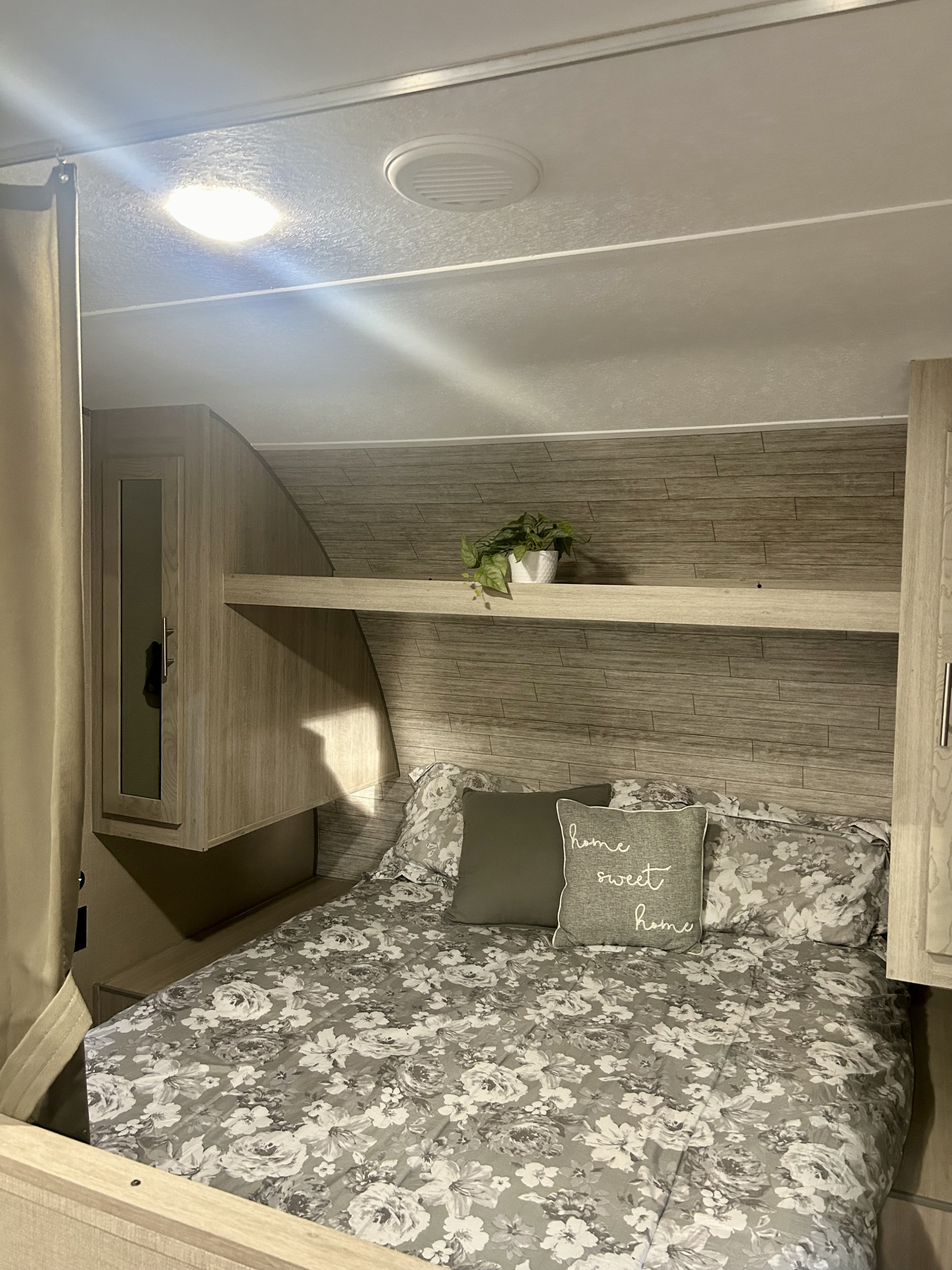 A cozy RV rental bedroom is shown with gray floral bedding, a "home sweet home" pillow, and overhead wood paneling with a shelf and plant.. Forest River Coachmen Catalina Summit 2023