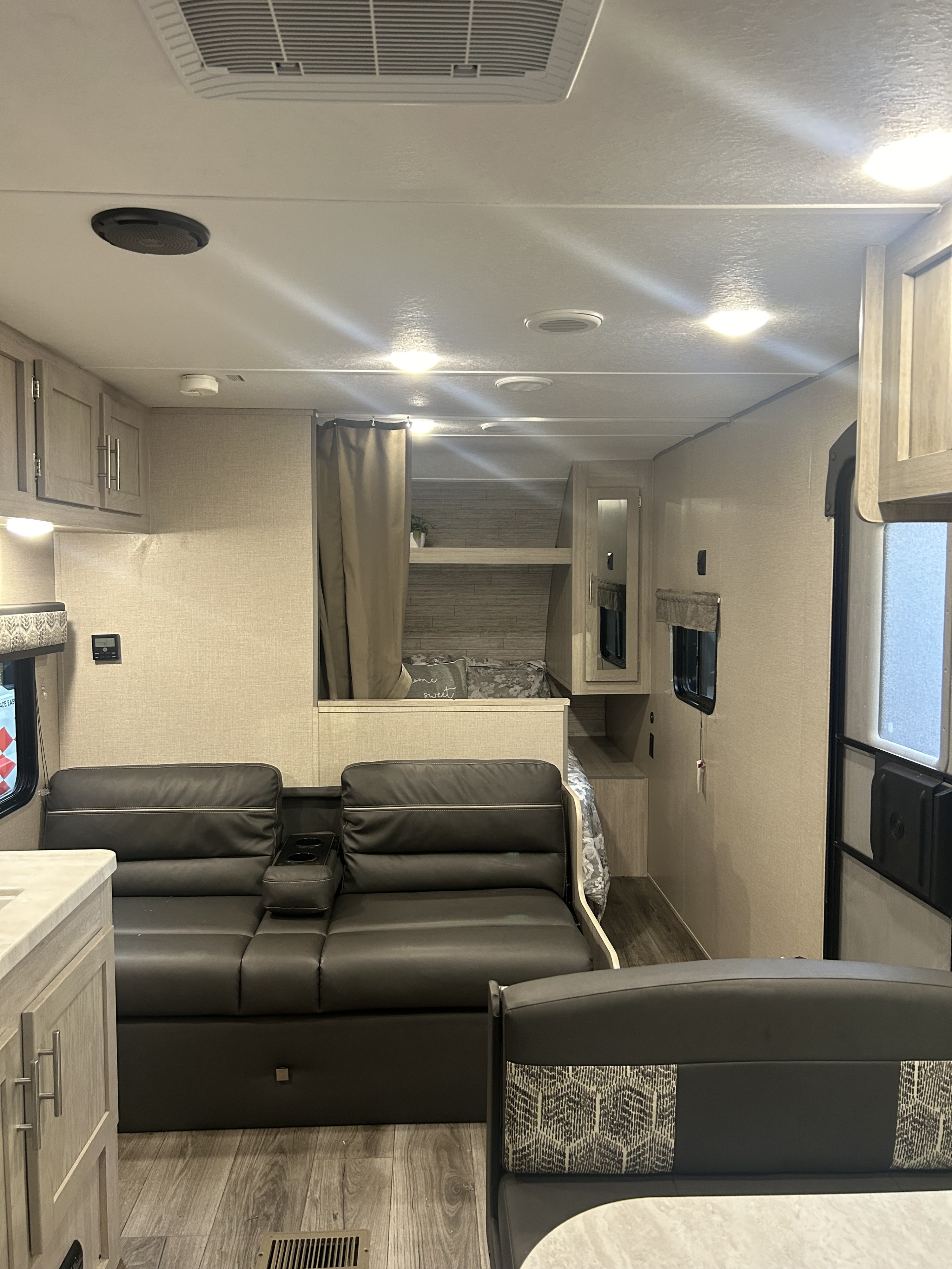 RV rental interior: living area with dark gray sofa, bed behind curtain, light wood finishes, and dinette seating.. Forest River Coachmen Catalina Summit 2023