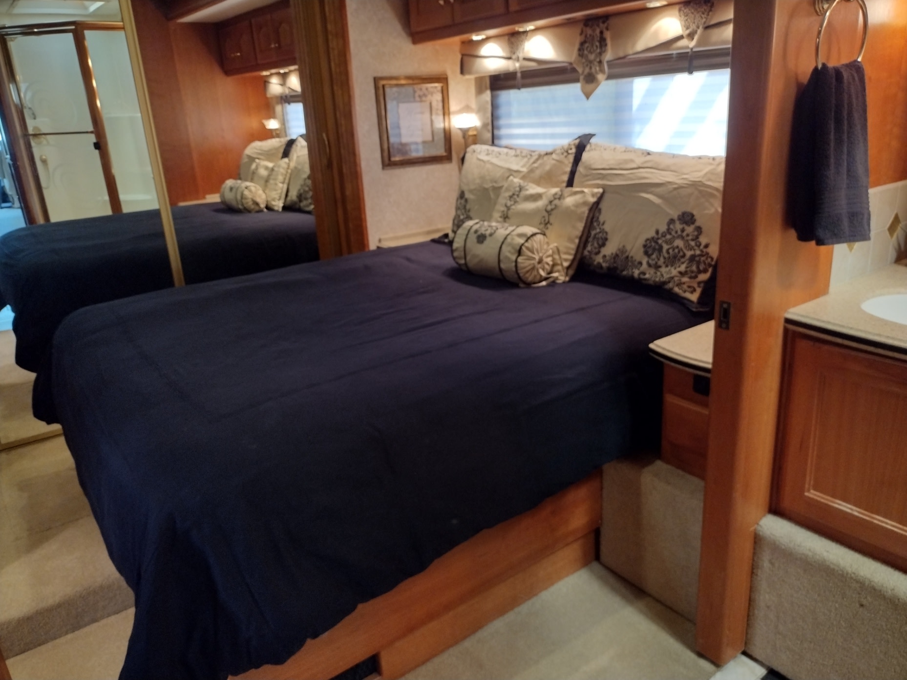 Cozy RV rental bedroom with a dark blue bed, decorative pillows, wood cabinetry. A mirrored wall and small sink area complete this motorhome interior.. Country Coach Allure 470 2005
