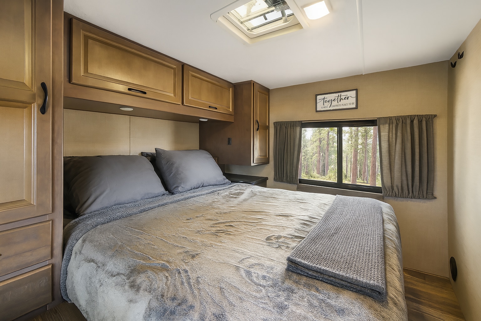 Cozy RV rental bedroom with soft grey bedding, overhead wood cabinets, and a scenic window view of a forest.. Thor Majestic 2018