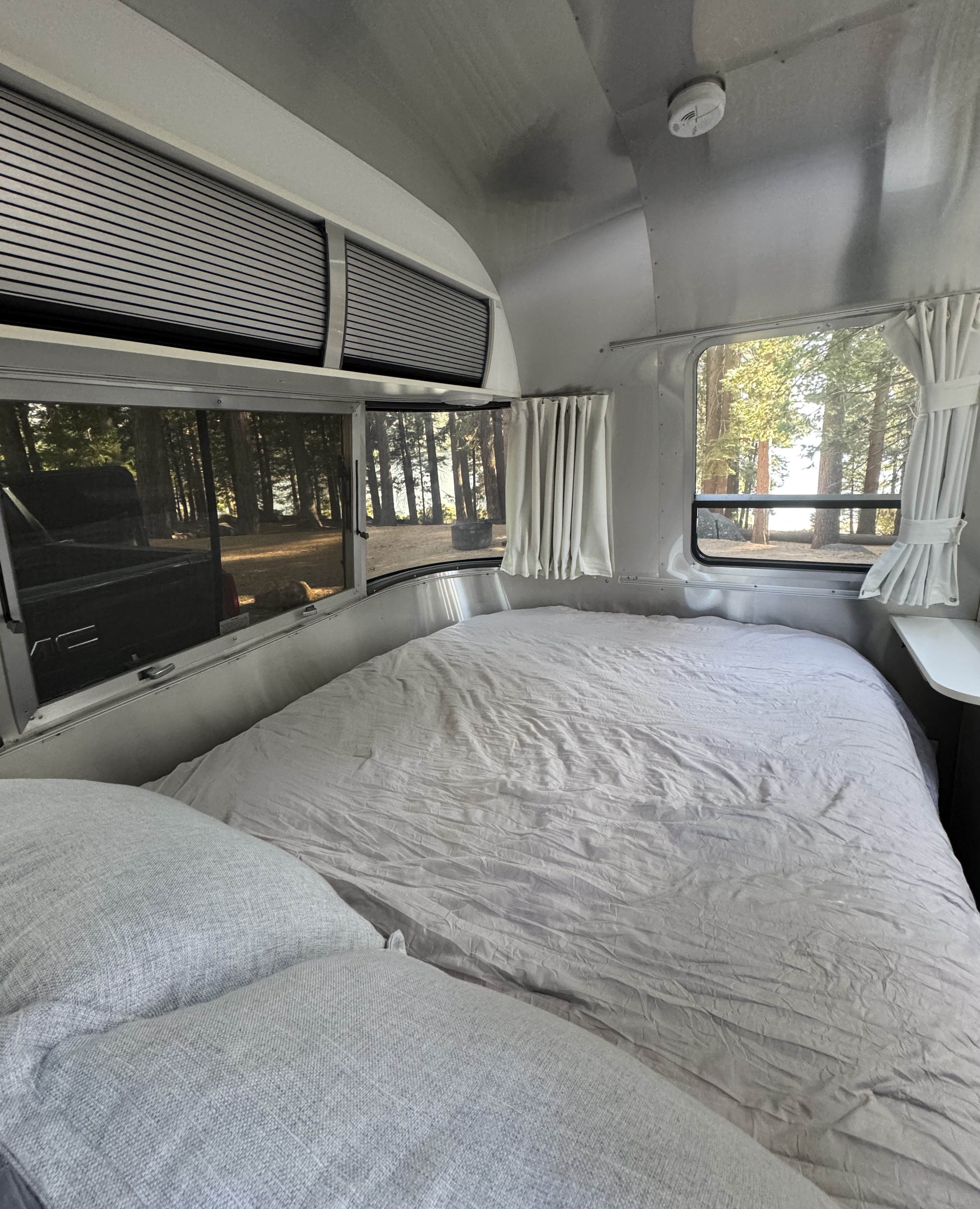 Cozy Airstream RV rental bedroom: light bedding, pillows, and large windows revealing a forest and distant water.. Airstream Sport 2018