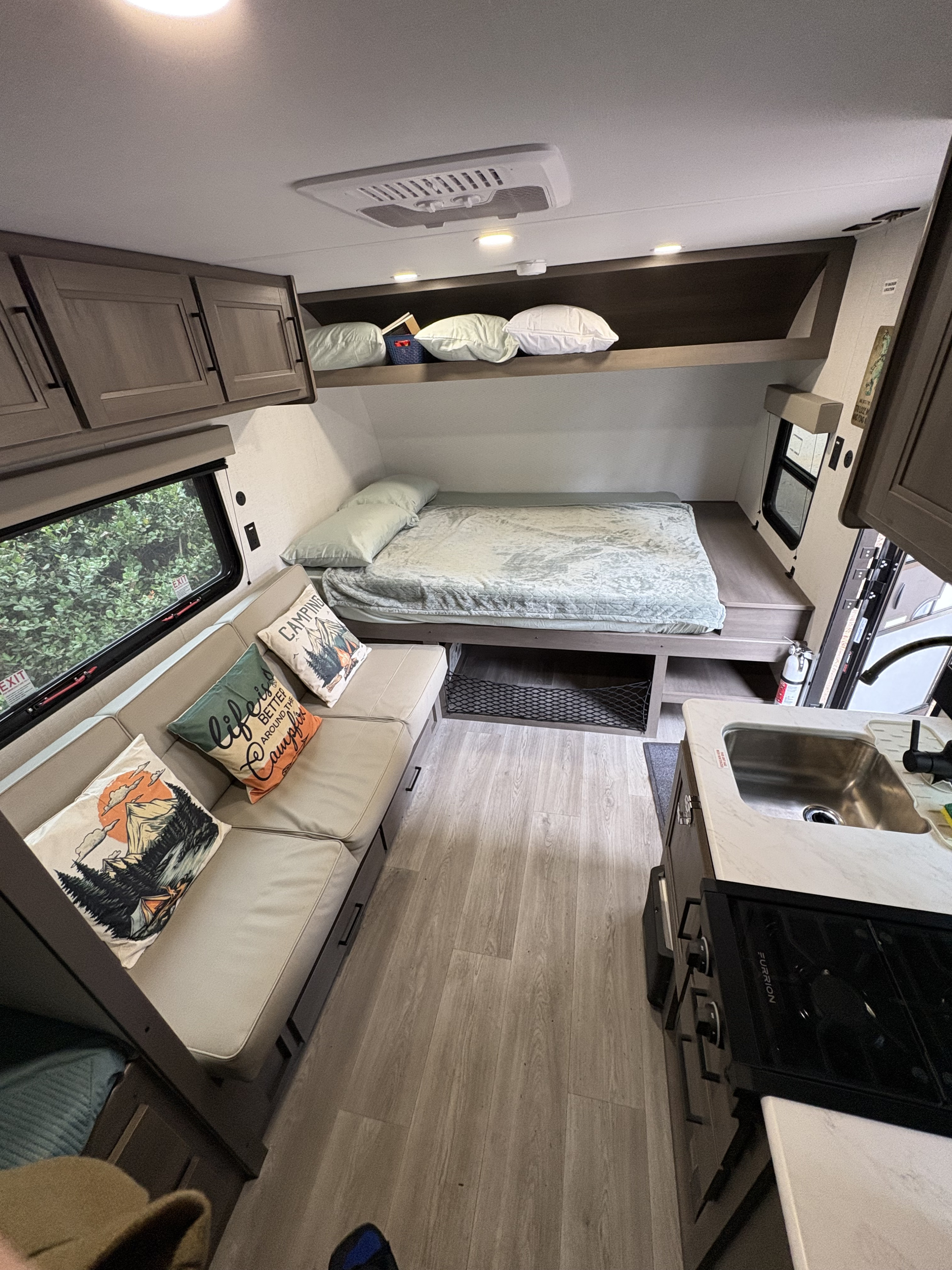 An RV rental interior with a comfy bed, overhead storage, sofa seating, a kitchen with a sink & stovetop, and wood-look floor.. Grand design 151bh 2025