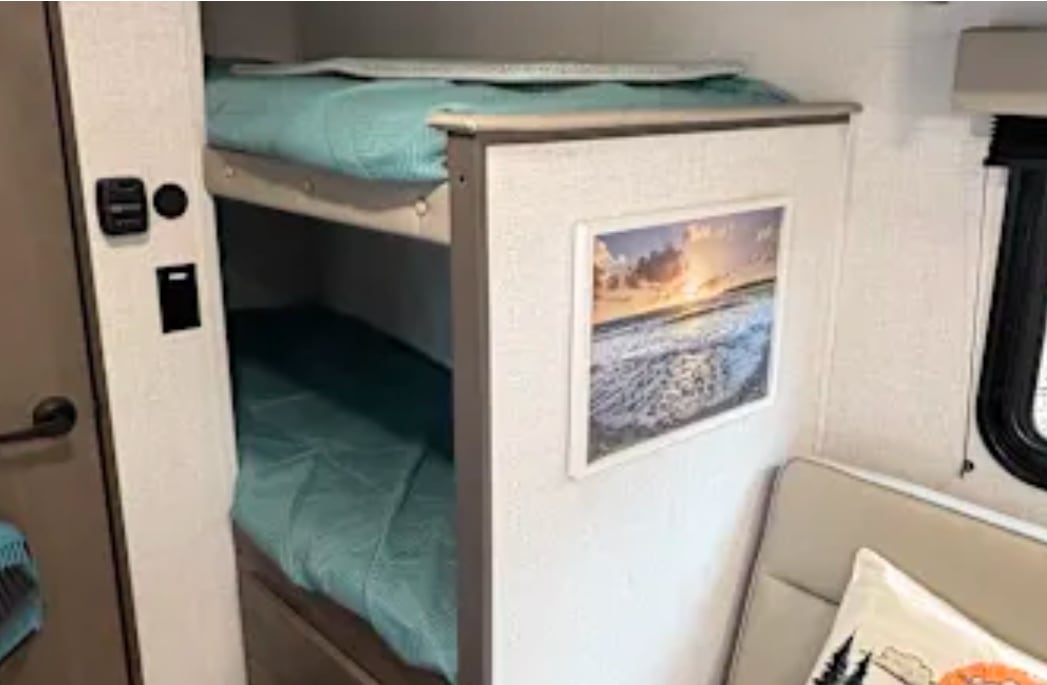 Cozy RV rental interior with inviting bunk beds featuring teal bedding. A beach scene print and cushioned seating add comfort.. Grand design 151bh 2025
