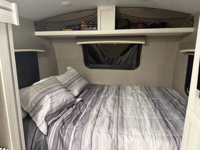 Cozy RV rental bedroom features a striped bed, overhead storage net with gear, and window with a shade, perfect for compact travel.. Forest River Rockwood Geo Pro 2021