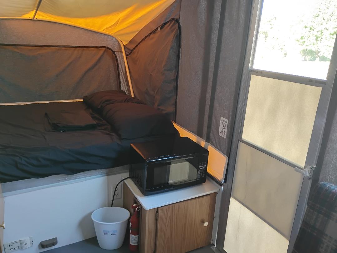 RV rental pop-up camper interior: black bed, microwave, fire extinguisher, and bucket next to a bright door.. Coleman Destiny Taos 1999