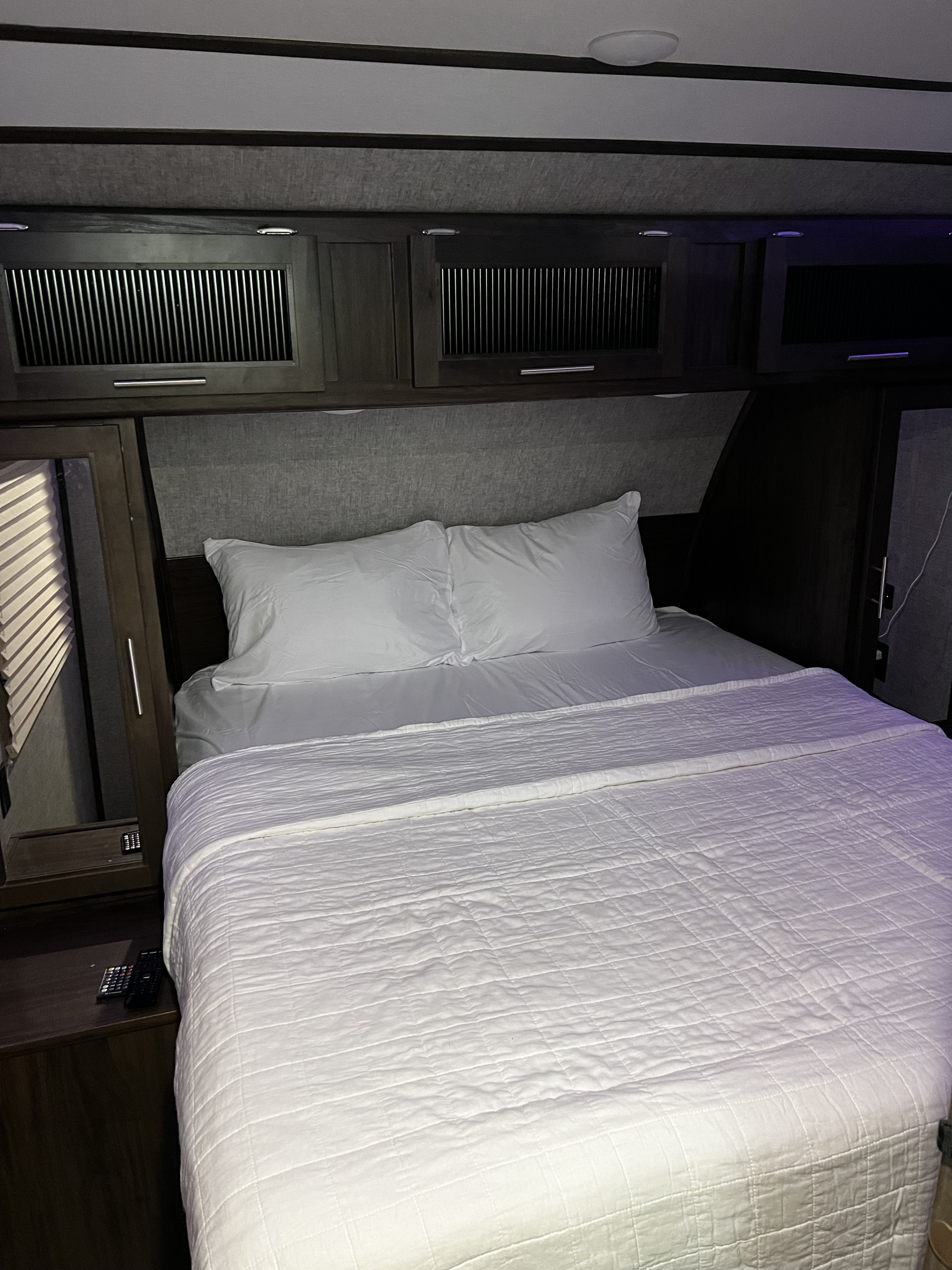 RV rental bedroom: A neatly made bed with white linens, pillows, and a quilted comforter under dark overhead cabinets.. Forest River Cherokee Alpha Wolf 2019