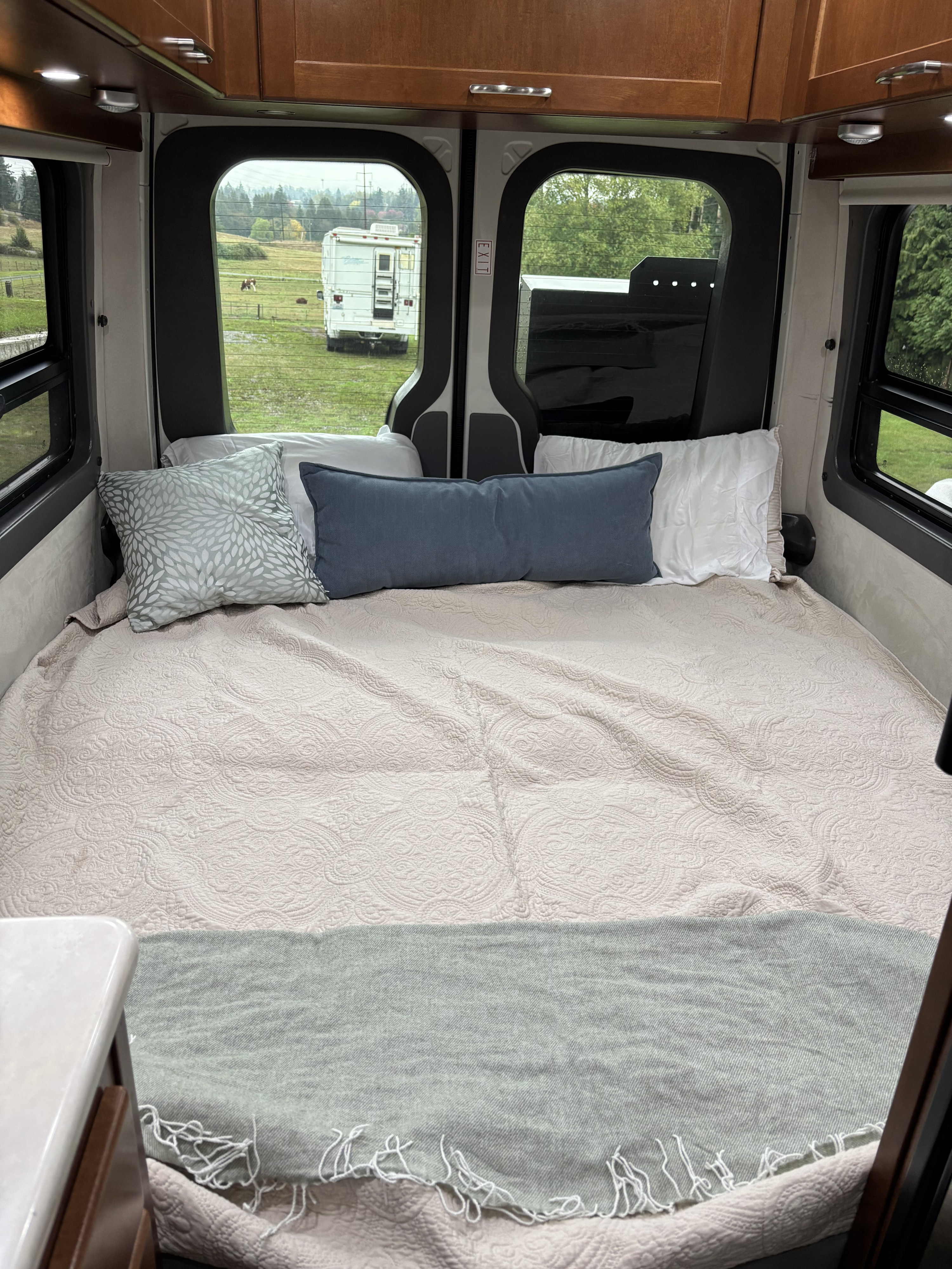 Cozy camper van bed for RV rental, featuring soft quilt, pillows, and serene views of a field and distant RV.. Pleasure-Way Pleasure-Way Class B 2016