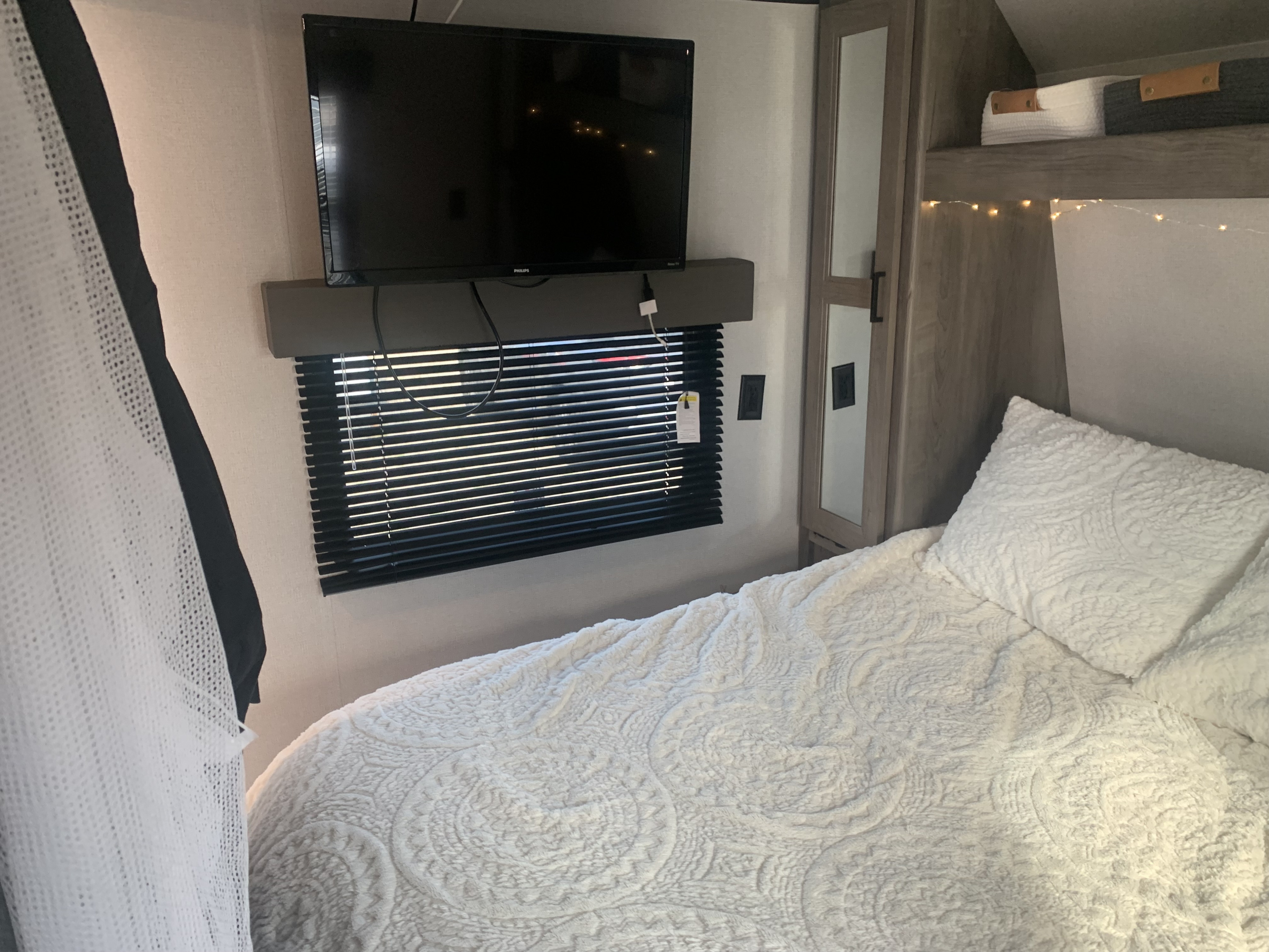 Cozy RV rental bedroom featuring a mounted TV, window with blinds, comfy white bed, mirrored wardrobe, and string lights.. Keystone RV Hideout 2021