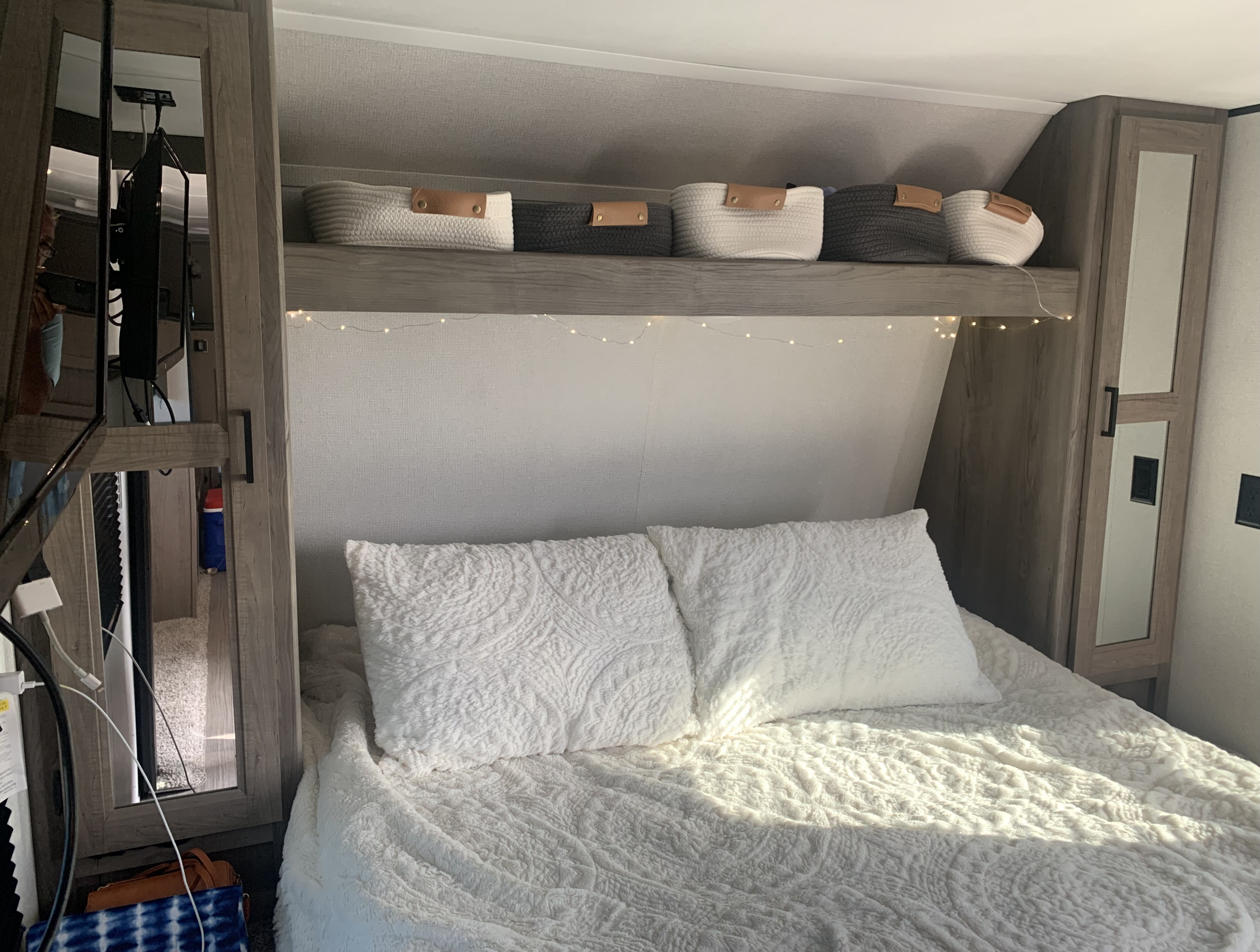 Cozy RV rental bedroom interior with a comfy white bed, woven baskets on a shelf, and warm fairy lights.. Keystone RV Hideout 2021