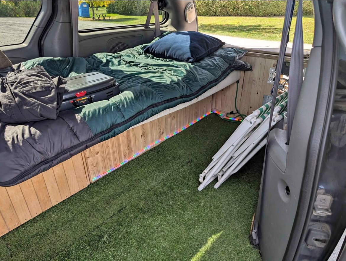 Inside an RV rental minivan conversion with a comfy bed, camp stove, artificial turf floor, and camping chairs.. Dodge Grand Caravan Camper 2004