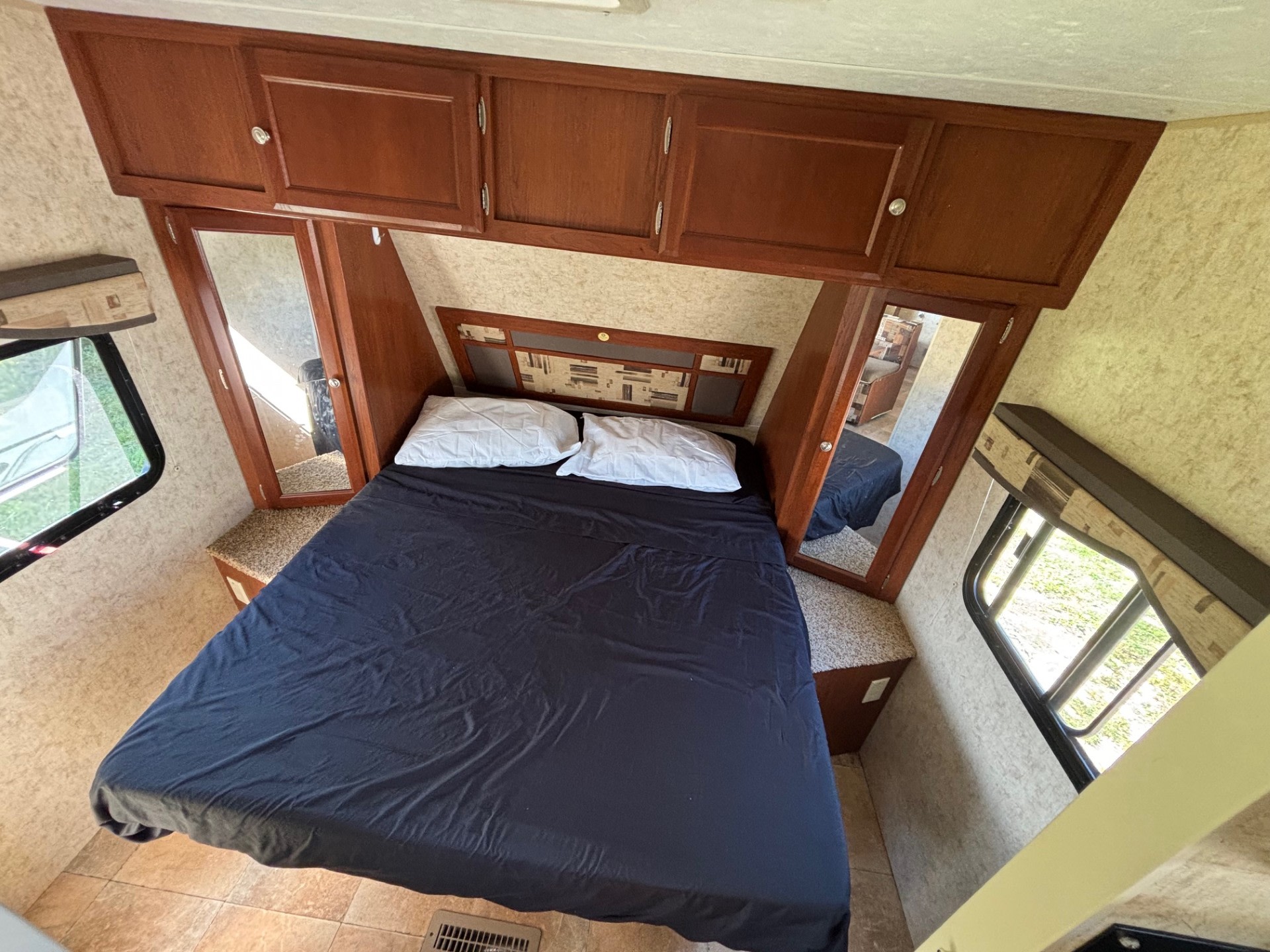 Inside an RV rental bedroom: a dark blue bed with white pillows, wood cabinets above, and side windows with green views.. Dutchmen Kodiak Ultra Lite 2011