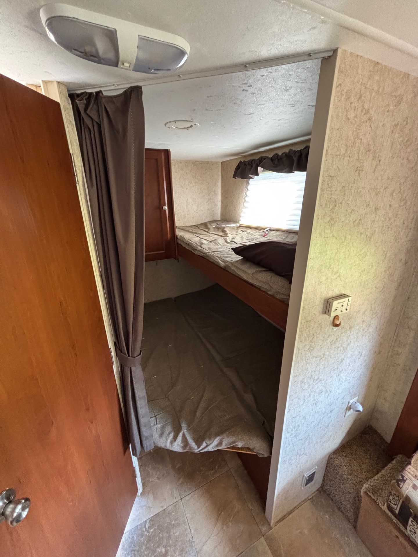 Interior of an RV rental showing a narrow bunk bed area with brown curtains, window, and a ceiling light.. Dutchmen Kodiak Ultra Lite 2011
