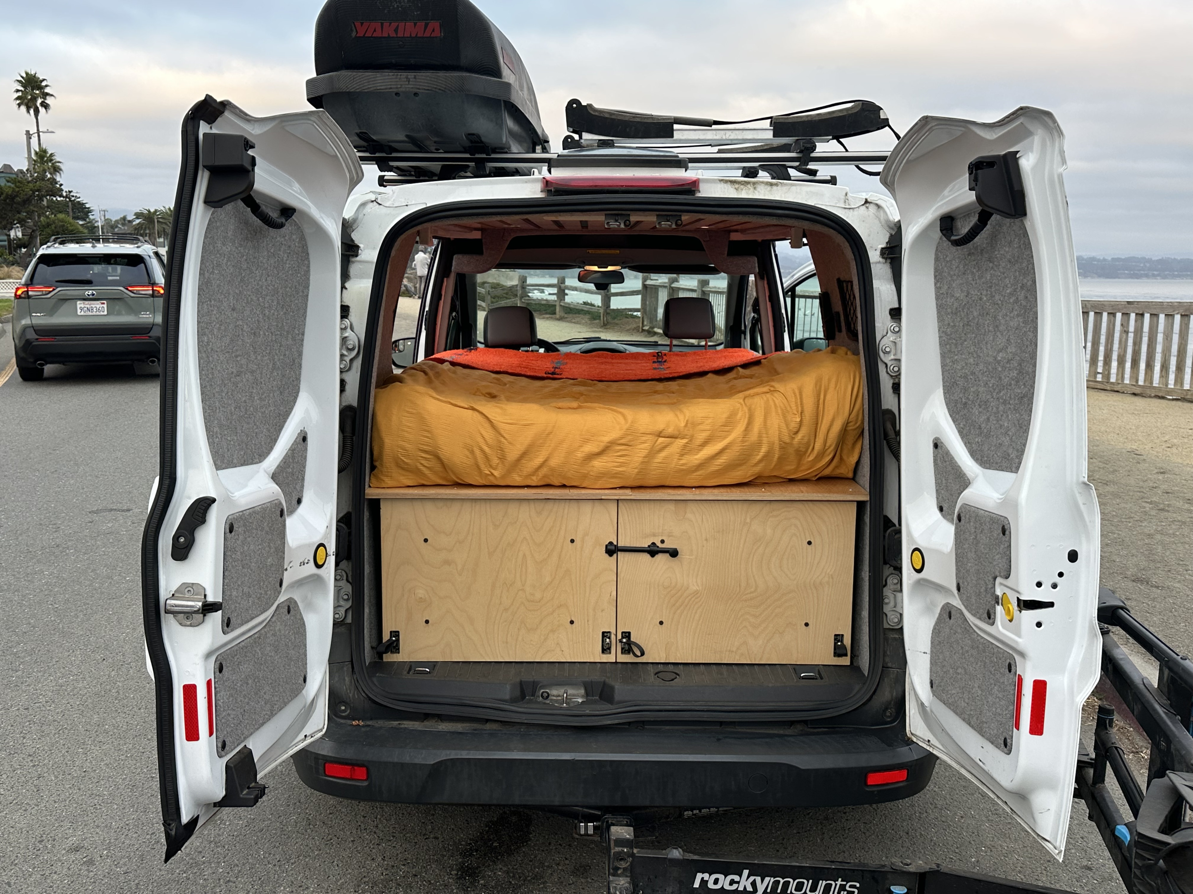 Rear view of an **rv rental** camper van. Open doors show custom bed/storage setup inside, Yakima roof box & board rack up top, by the coast.. Ford Transit custom 2018