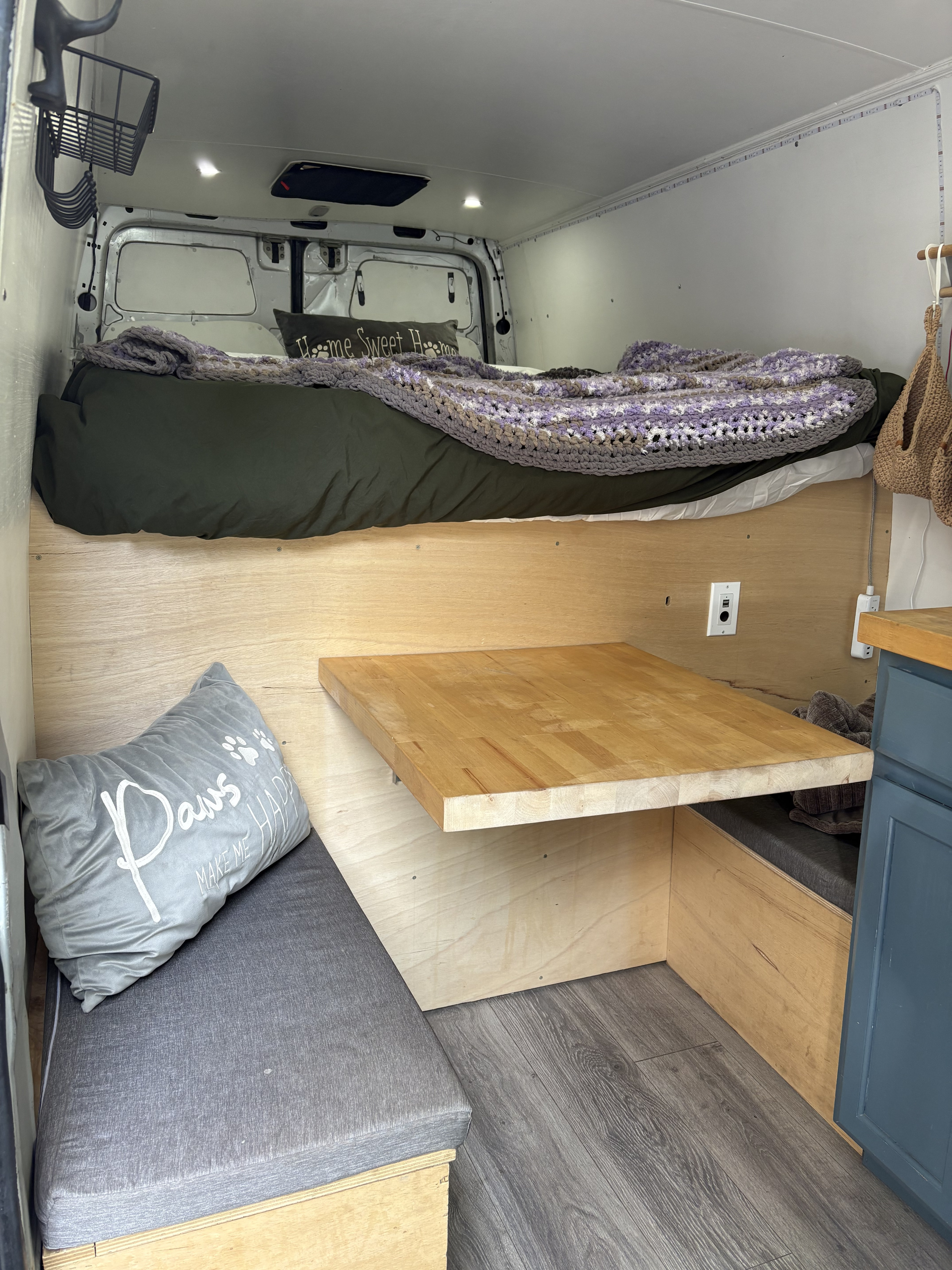 This cozy rv rental camper van interior features a raised bed, fold-out wooden table, and grey cushioned seating, perfect for compact travel.. Mercedes Sprinter 2014
