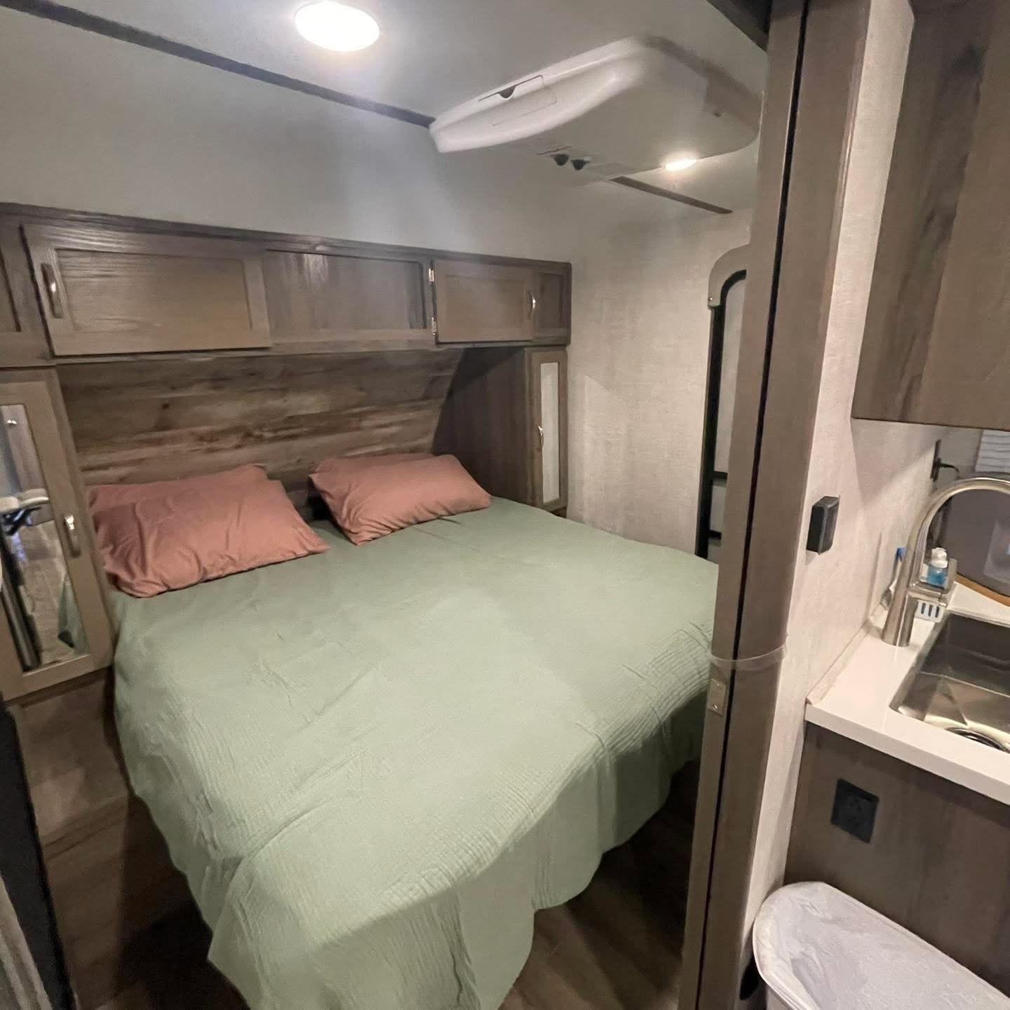 Cozy RV rental bedroom: bed with green comforter & pink pillows, wooden overhead cabinets, and AC unit overhead.. Gulf Stream Envision 2023