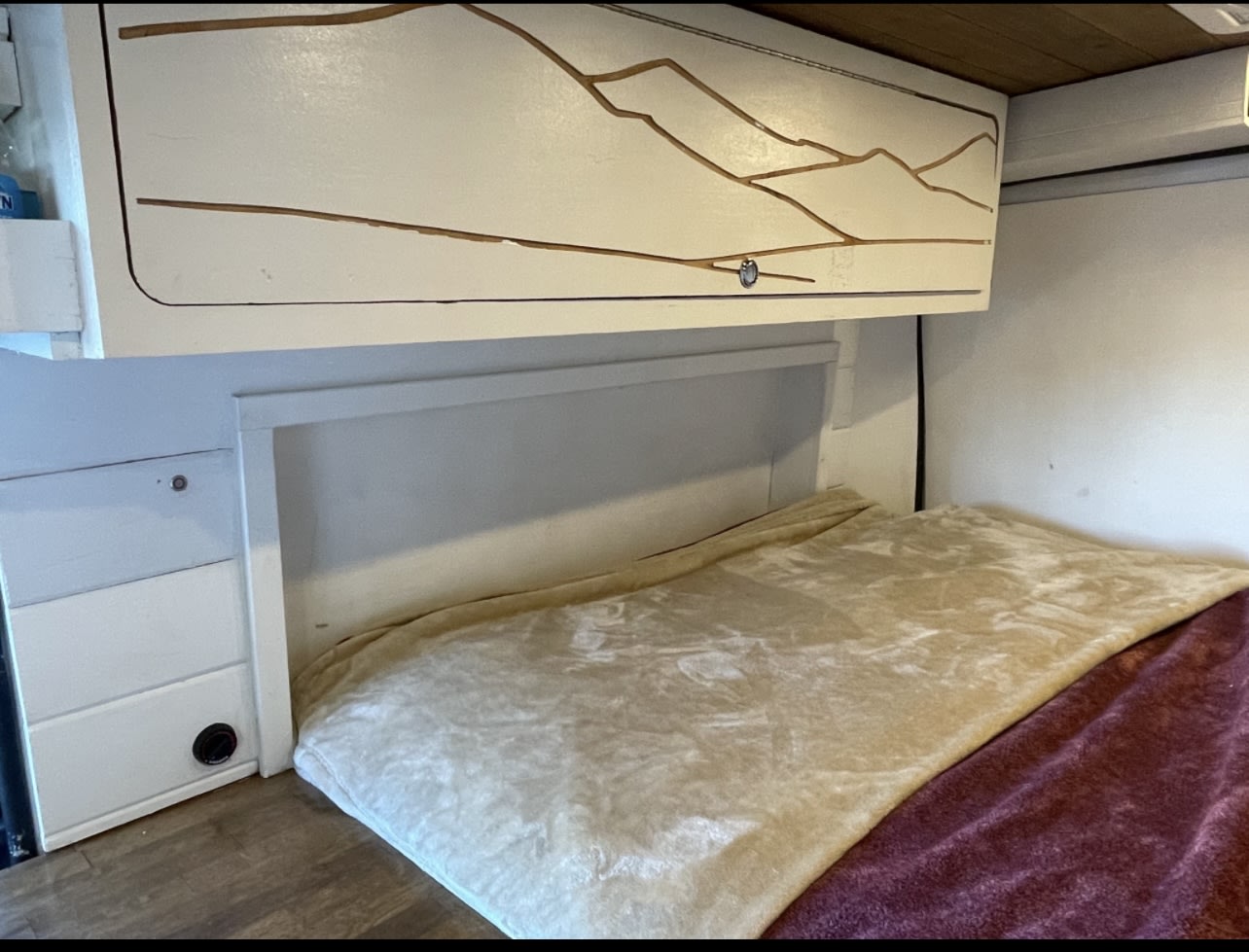 Cozy RV rental interior with bed, storage, and a mountain-themed overhead cabinet. Ideal for your next camper van trip.. Ram Promaster 2021