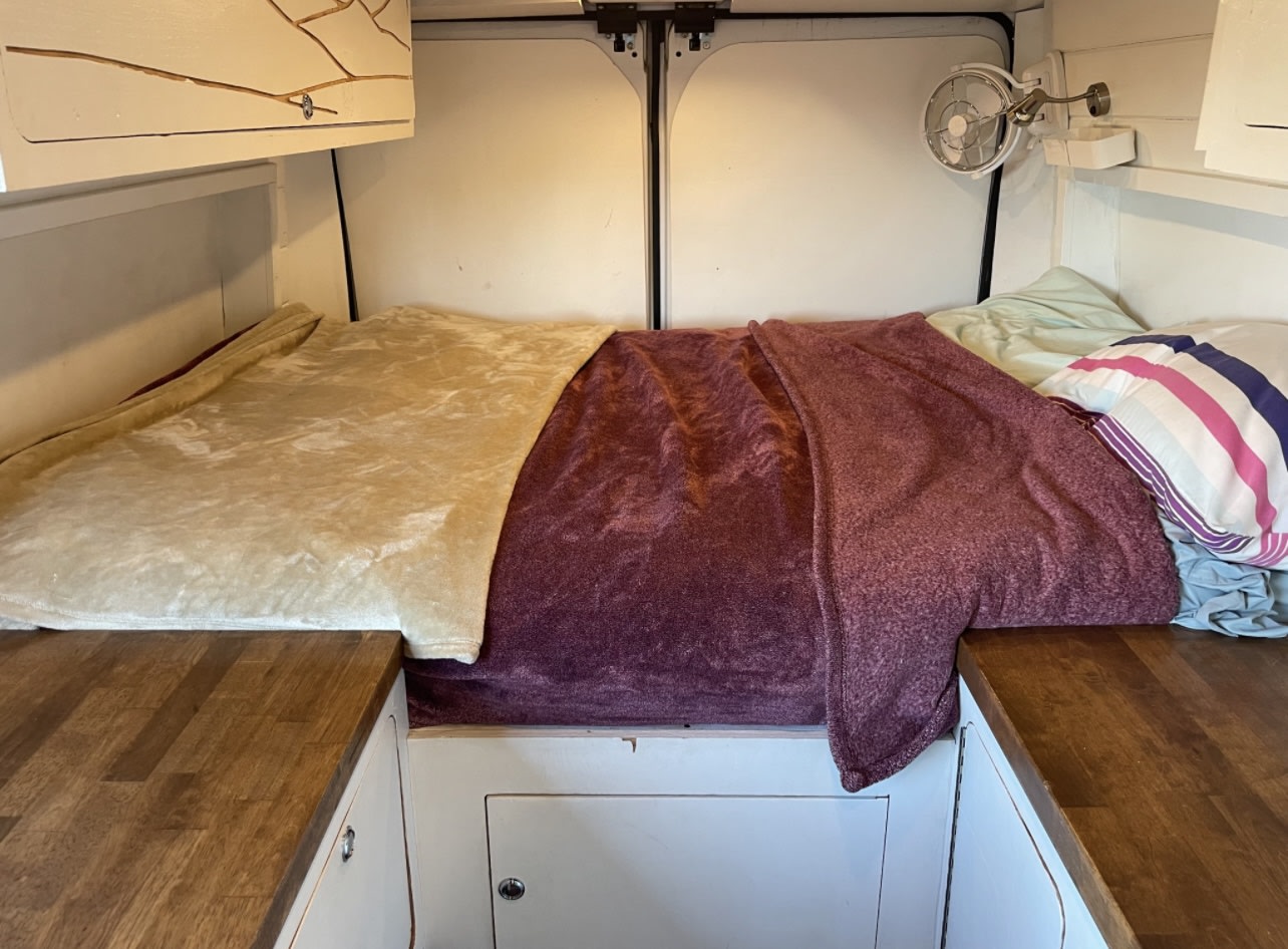 Comfortable custom RV rental interior with a bed, blankets, fan, and wood countertops, ideal for travel.. Ram Promaster 2021