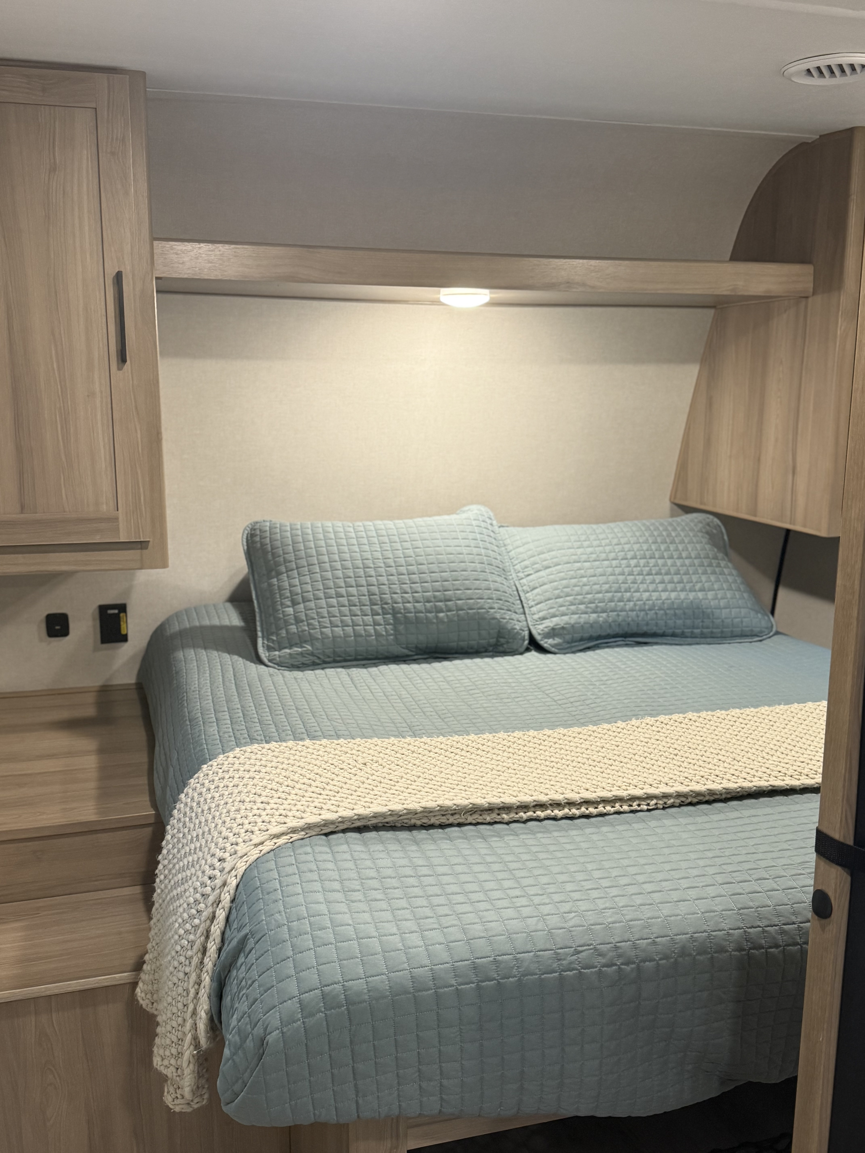 Cozy RV rental bedroom featuring a comfortable bed with blue quilted bedding, overhead shelf light, and wood cabinetry.. Keystone RV Keystone Coleman Light 22BX 2025
