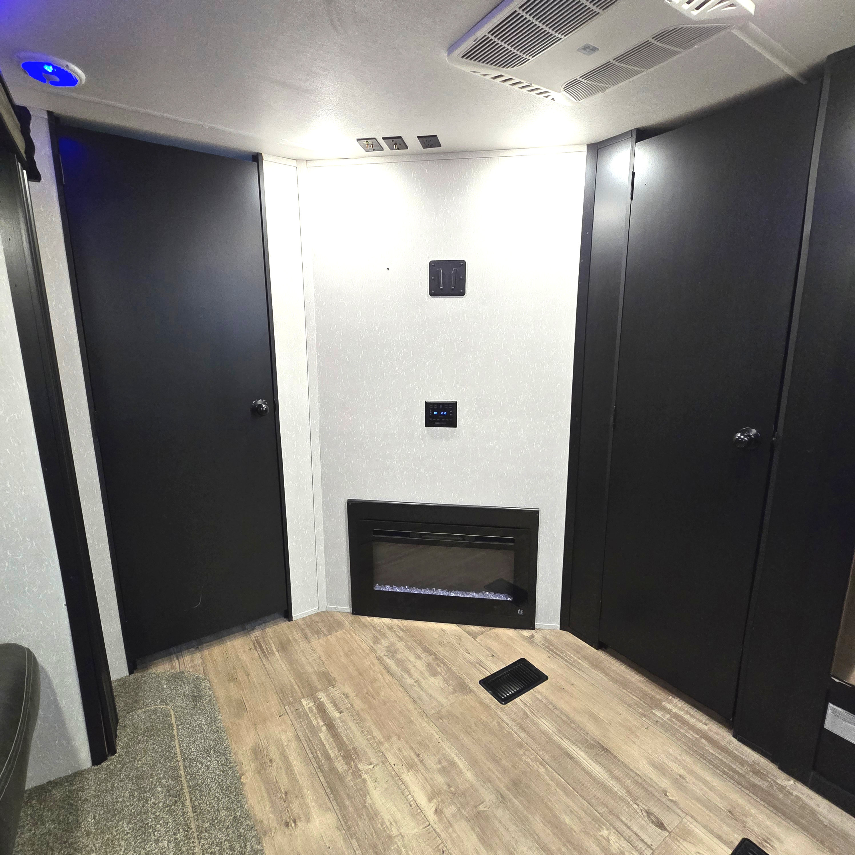 RV rental interior with a modern electric fireplace, two dark doors, TV mount, controls, wood floor, and ceiling AC/vents.. Forest River Cherokee Limited 2021
