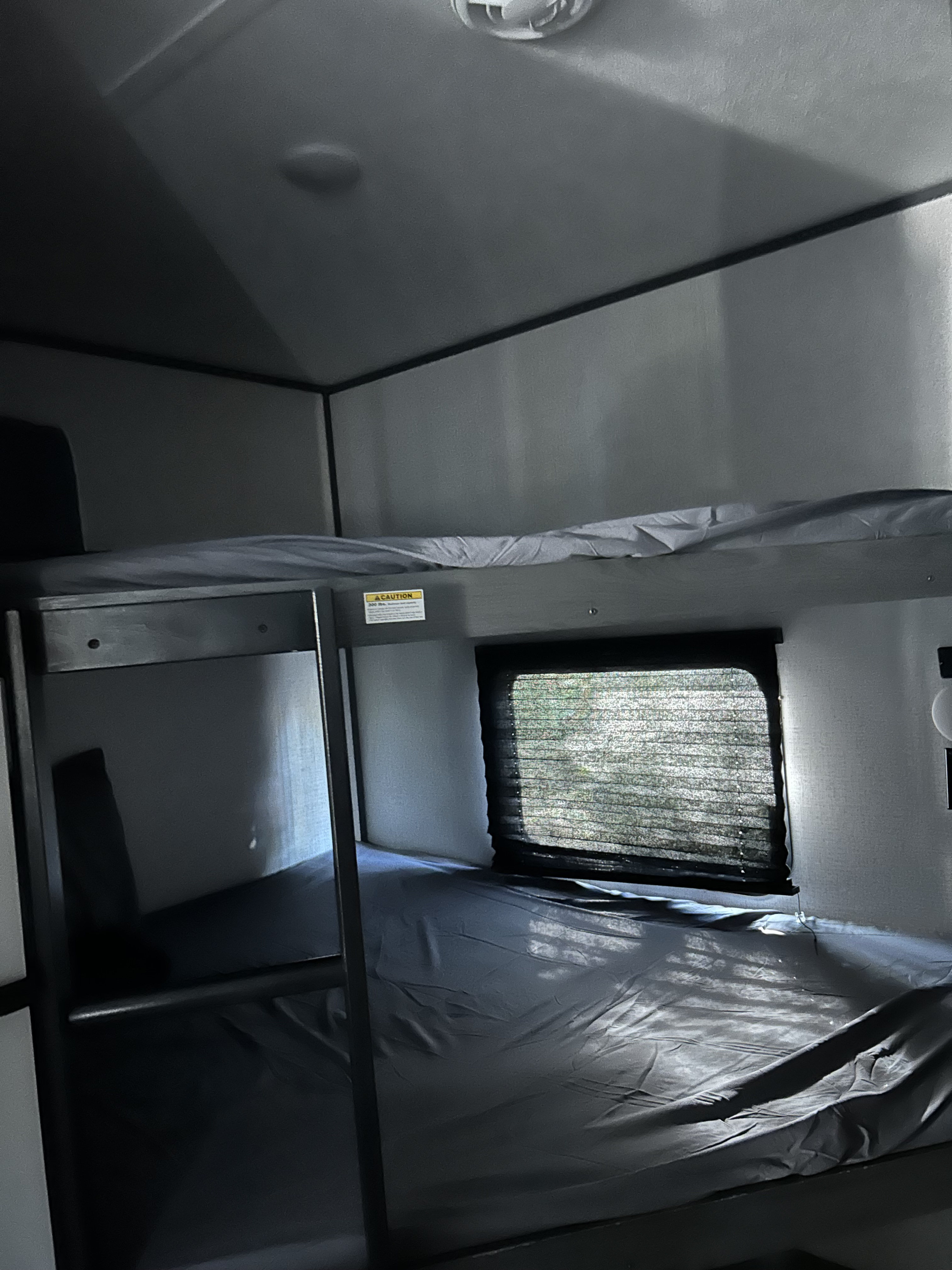 RV rental bunk beds, dimly lit with grey sheets. A caution sticker is visible, and a window shows outdoor greenery through blinds.. Dutchmen 3120BHS 2021