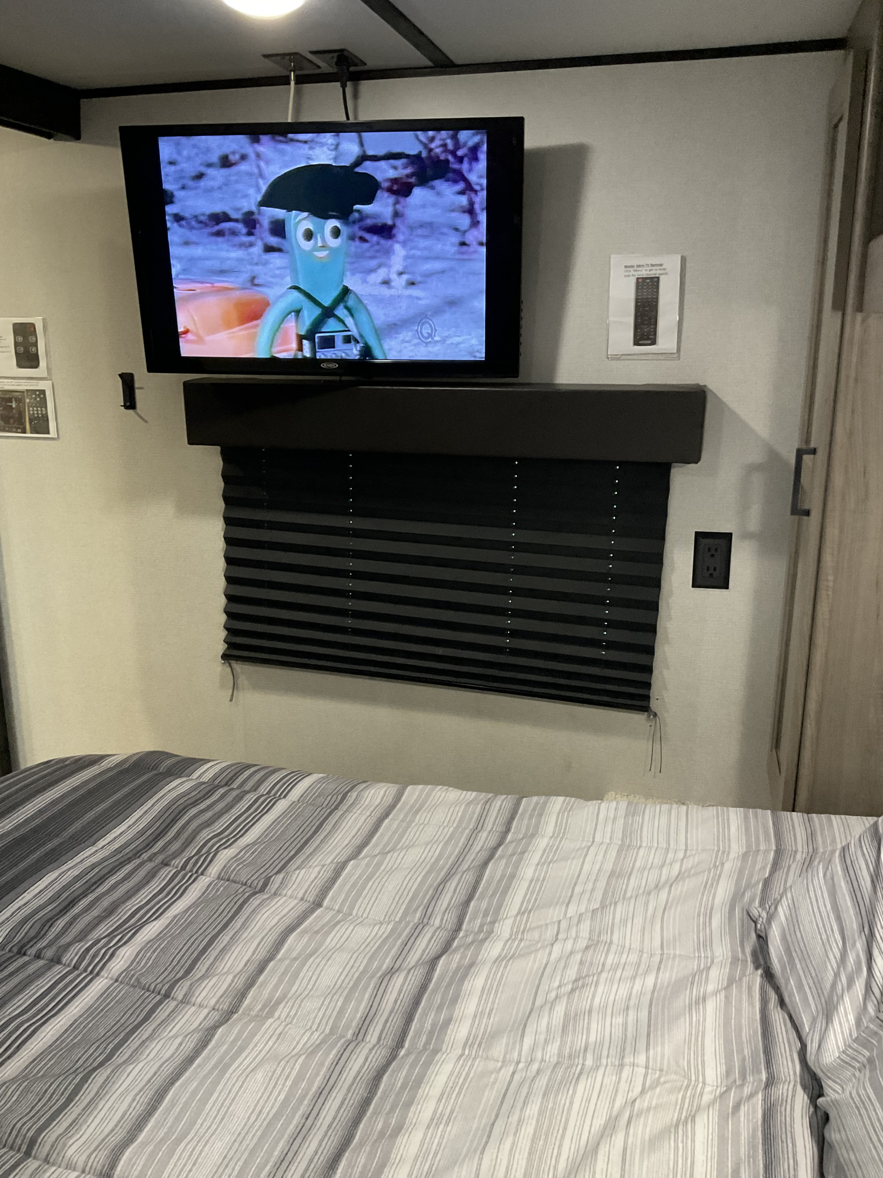 Cozy RV rental bedroom with TV playing Gumby, mounted above black blinds, and a striped bed in the foreground.. Keystone RV Hideout 2021
