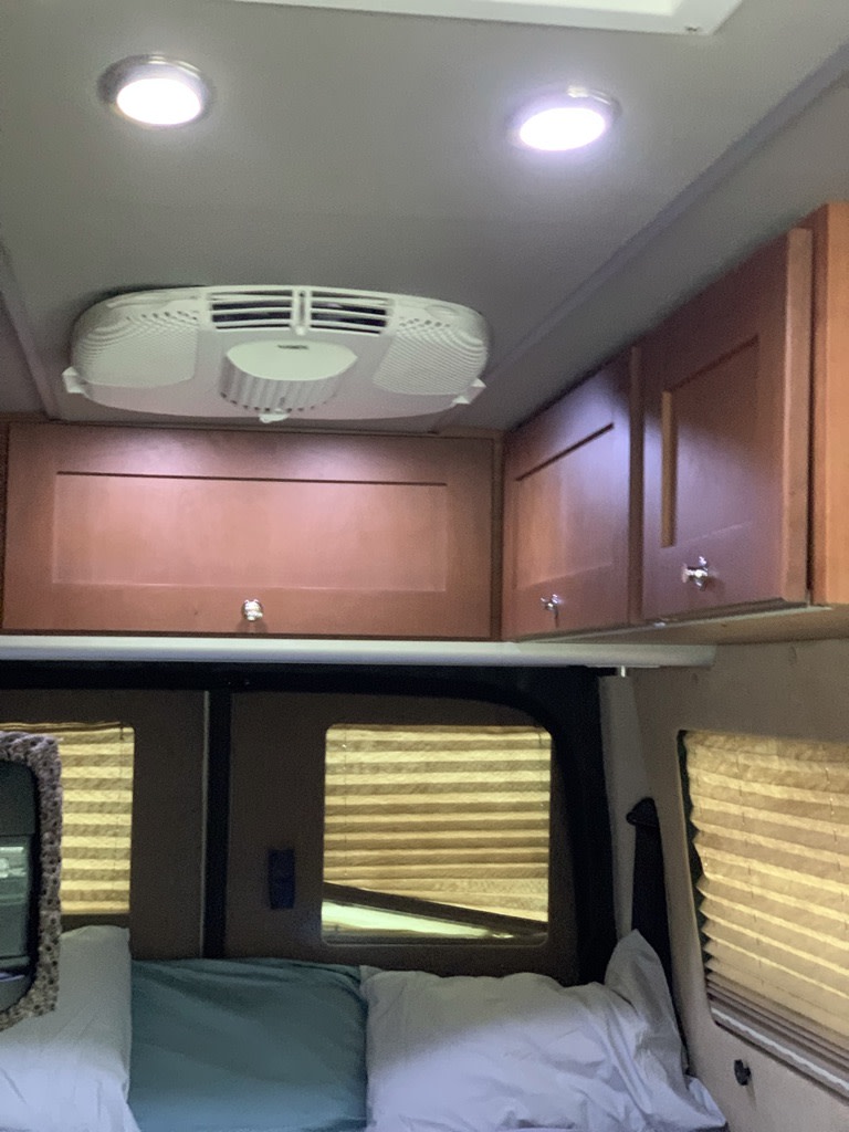 Inside an RV rental, showing the ceiling AC unit, bright lights, wooden cabinets, and a cozy sleeping area with windows and blinds.. Roadtrek ETREK 4x4 2019