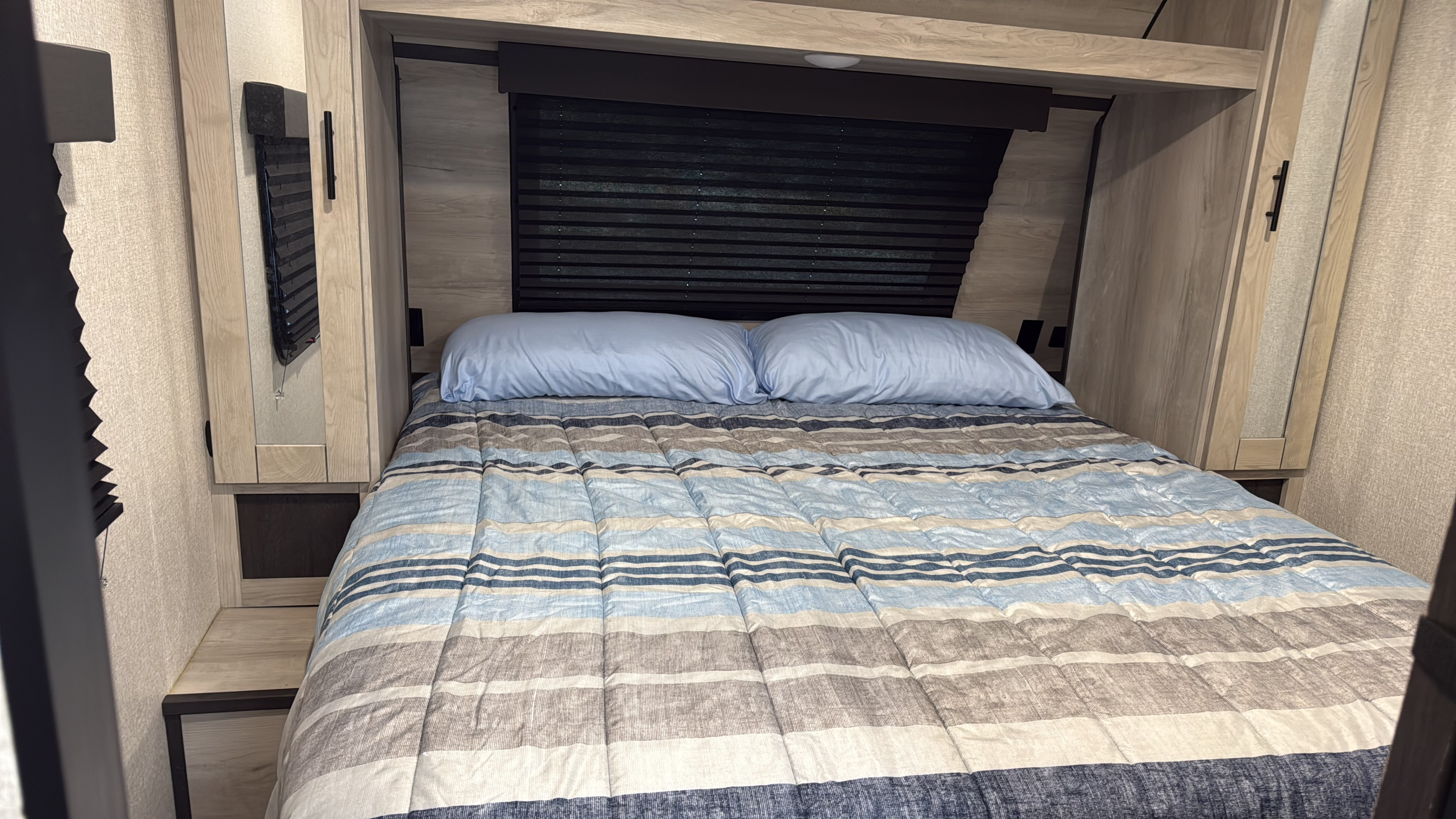 Cozy RV rental bedroom featuring a bed with striped comforter, blue pillows, wooden storage, and a privacy blind for relaxation.. KZ C261RB 2021