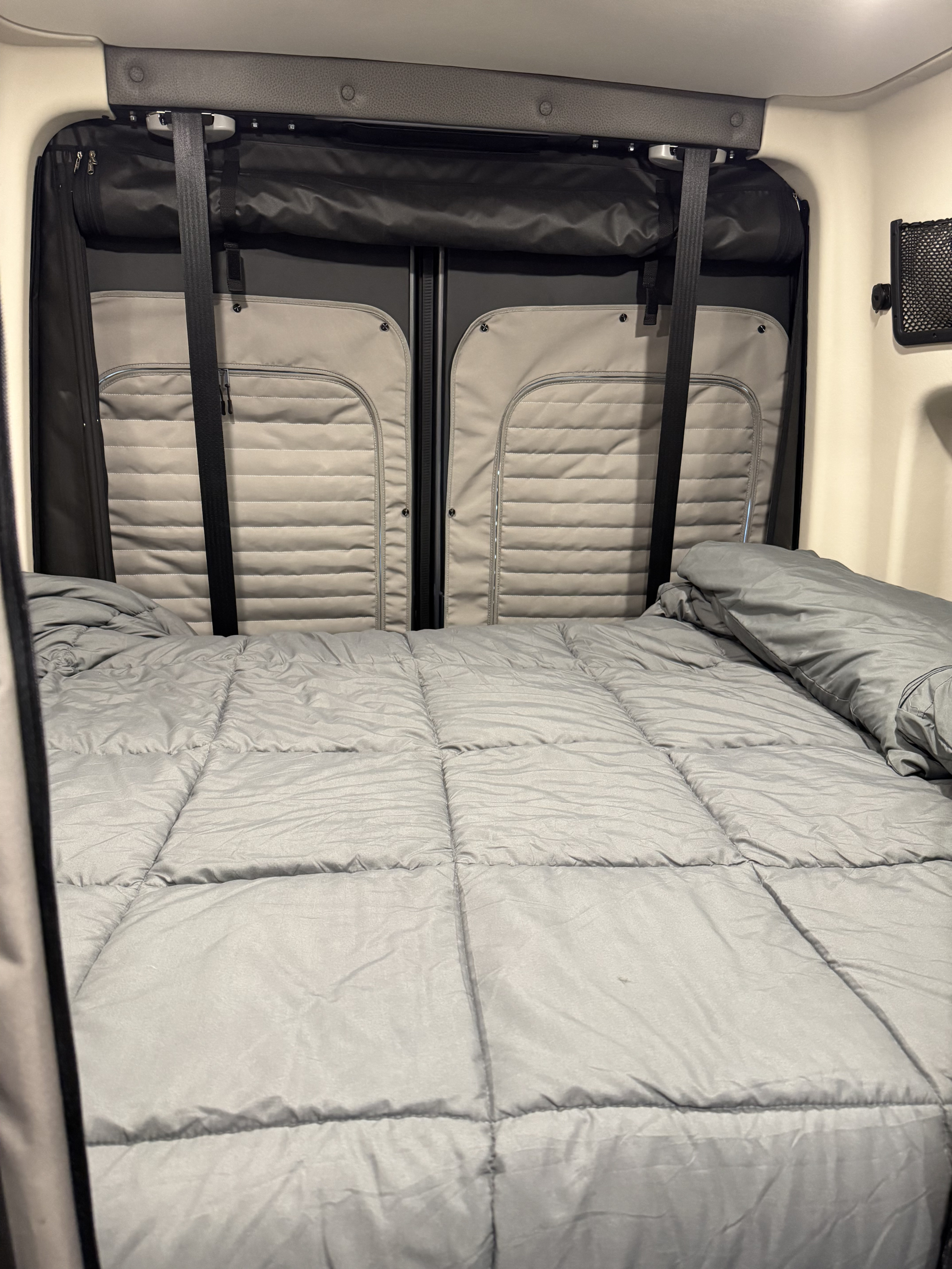 Comfortable sleeping area in an rv rental, featuring a bed with a grey comforter and padded rear door panels.. Winnebago Revel 2025