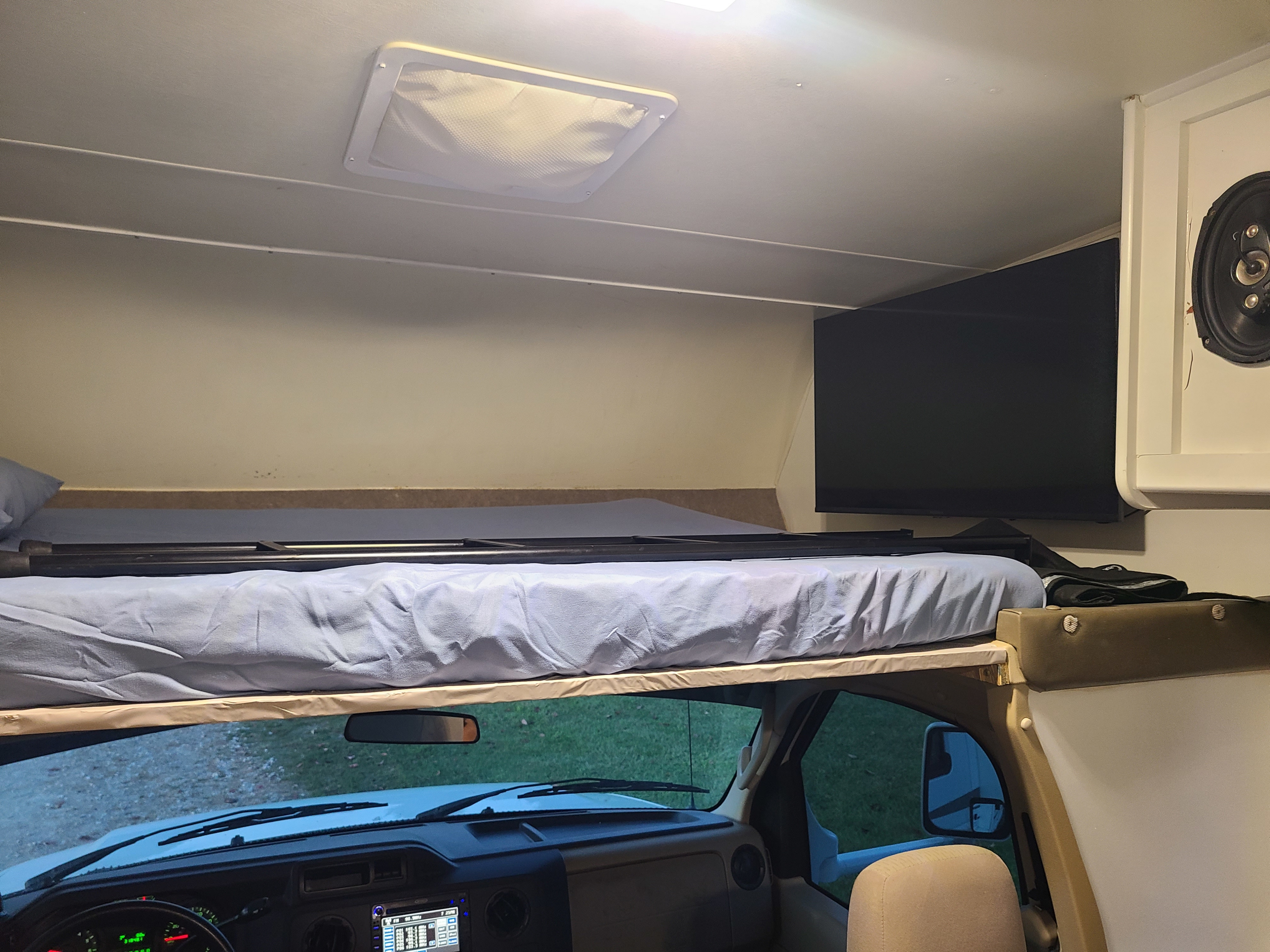 An RV rental interior features an over-cab bunk bed with blue bedding, a mounted TV, and front dashboard details below.. CoachMen Freelander 2012