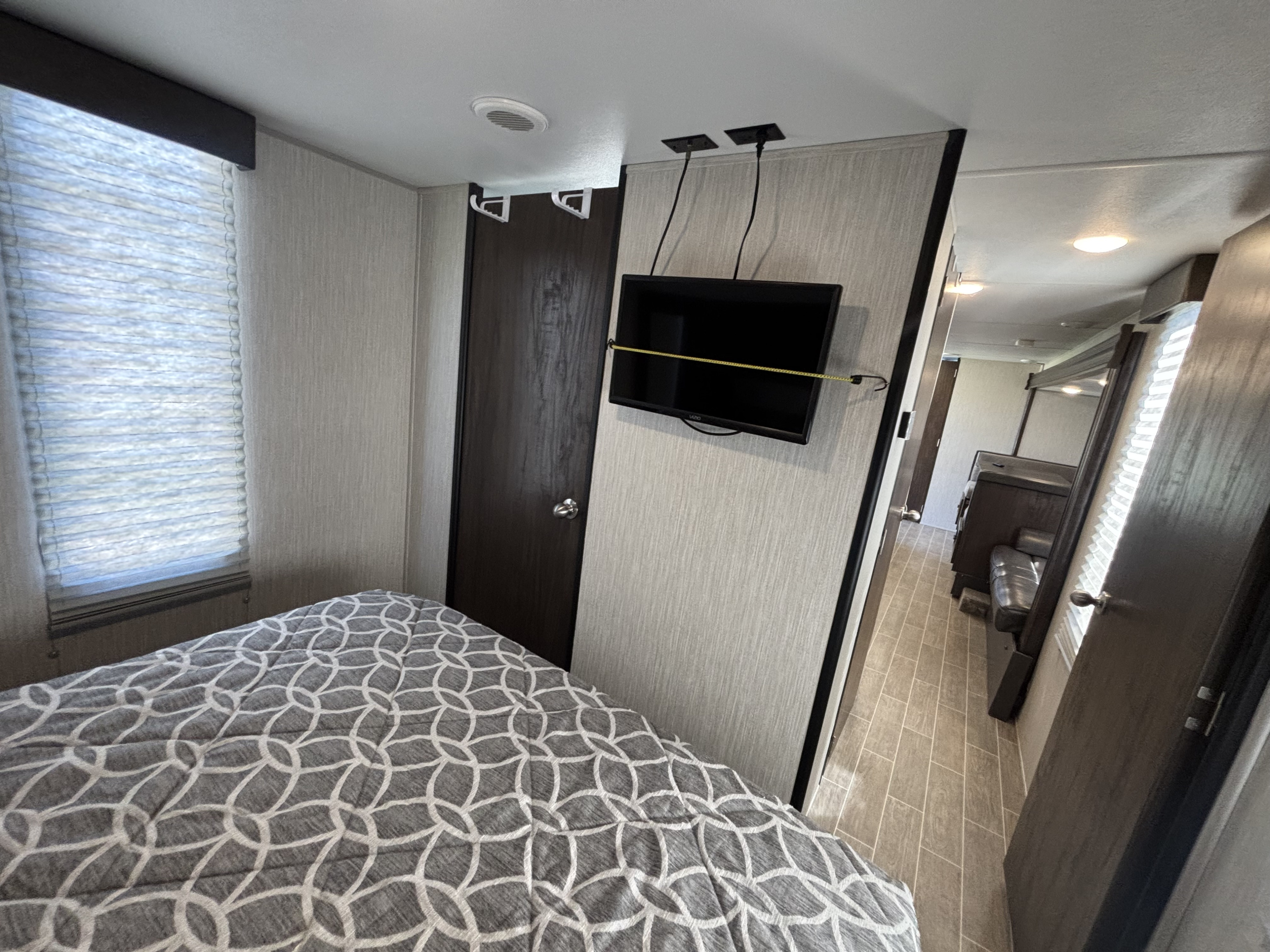This RV rental bedroom has a cozy patterned bed, wall-mounted TV, and window blinds, leading to the main living space.. Forest River Cherokee Grey Wolf 2020