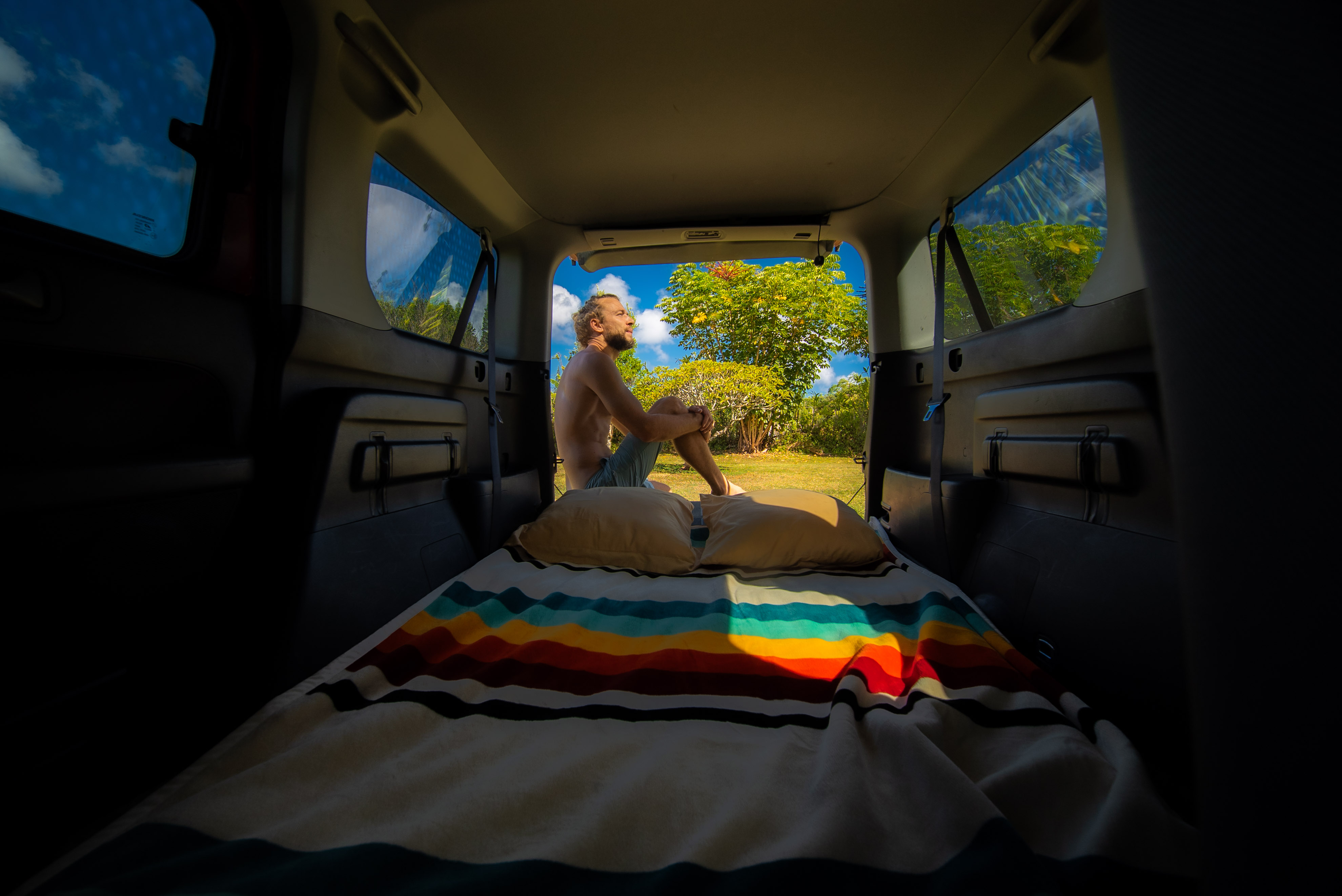 RV rental comfort: Man relaxes on his colorful, striped bed inside a van, gazing at lush trees and a blue sky through the open back.. Honda Element 2004