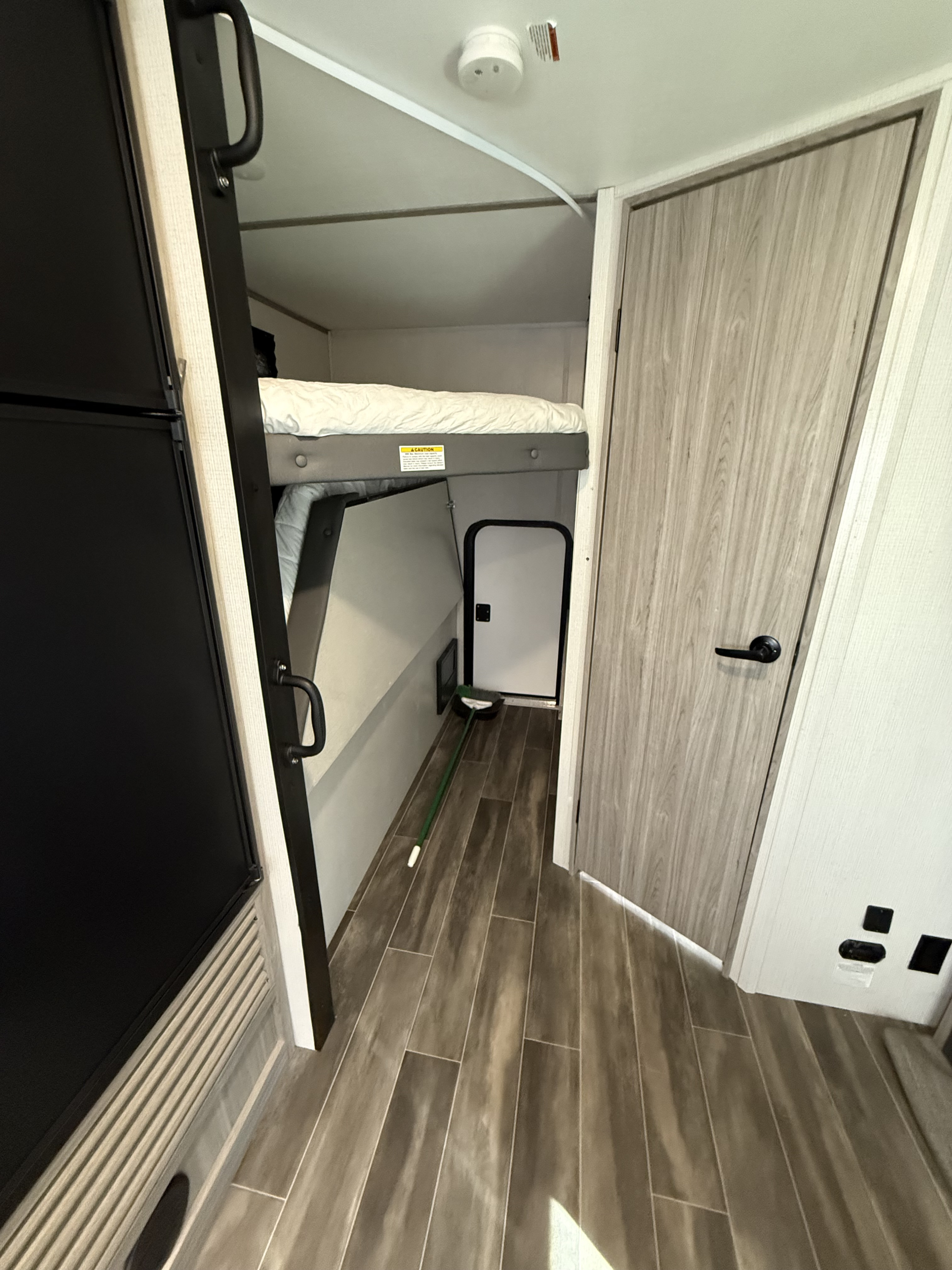 RV rental interior shows bunk beds, a refrigerator on the left, a doorway on the right, and a broom on the wood-look floor.. Keystone RV Passport SL 2022