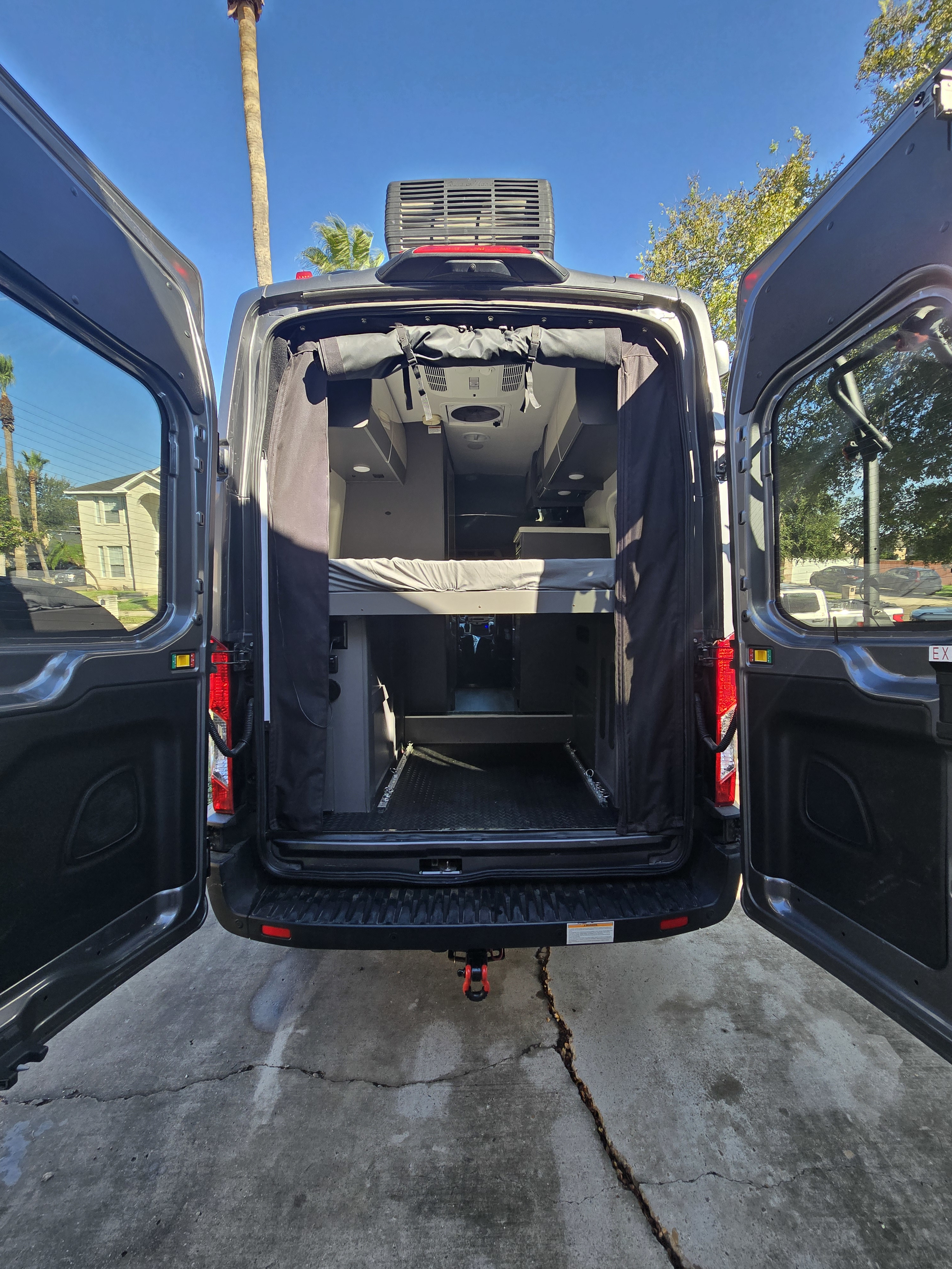 Open rear doors of a dark gray rv rental camper van reveal a bright interior with a raised bed, storage, and AC unit.. Thor Motor Coach Tranquility 19LT 2023