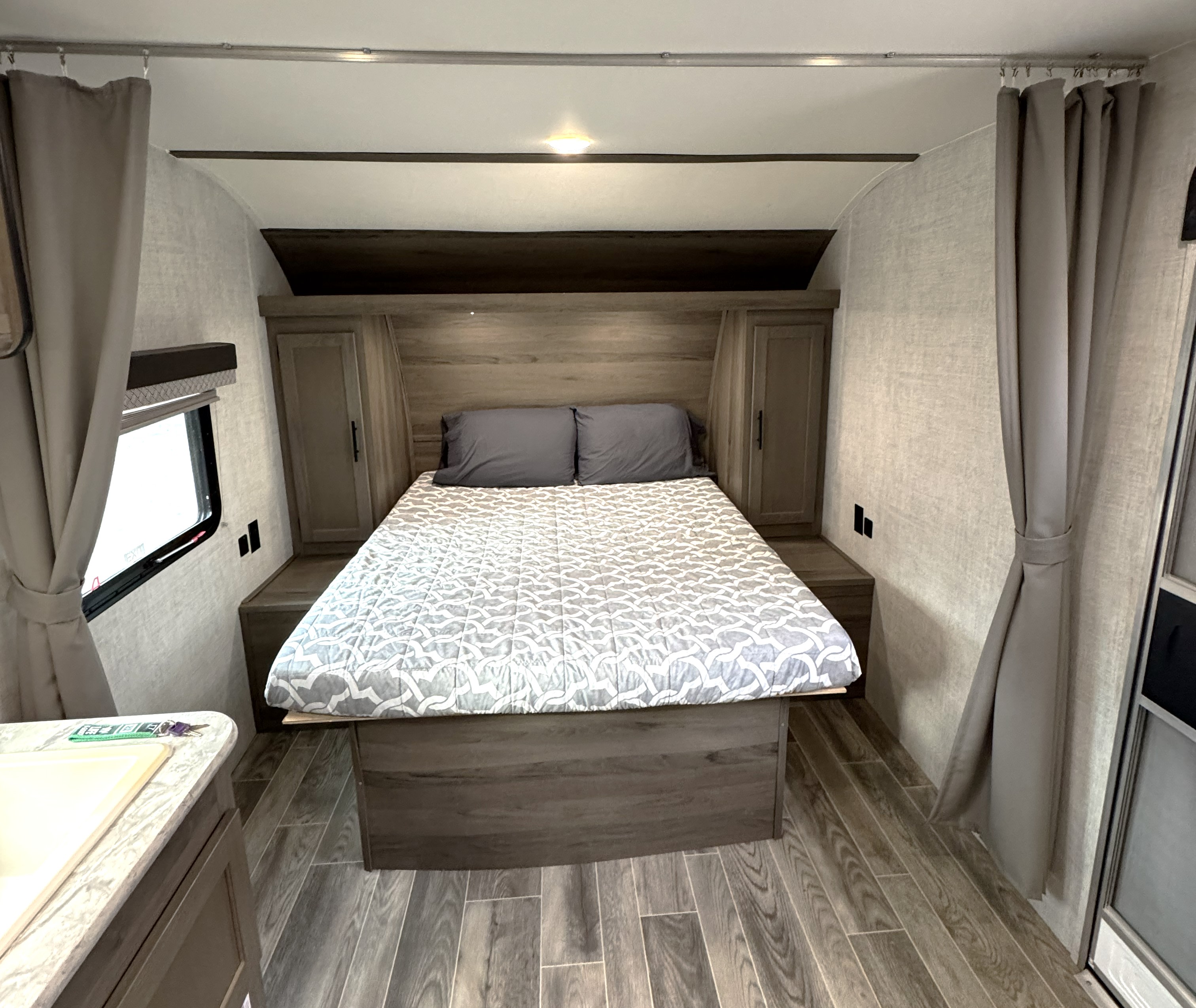 This cozy RV rental bedroom features a comfy bed, wood-look decor, side cabinets, and a window with curtains for a relaxing stay.. Gulf Stream Amerilite 2021