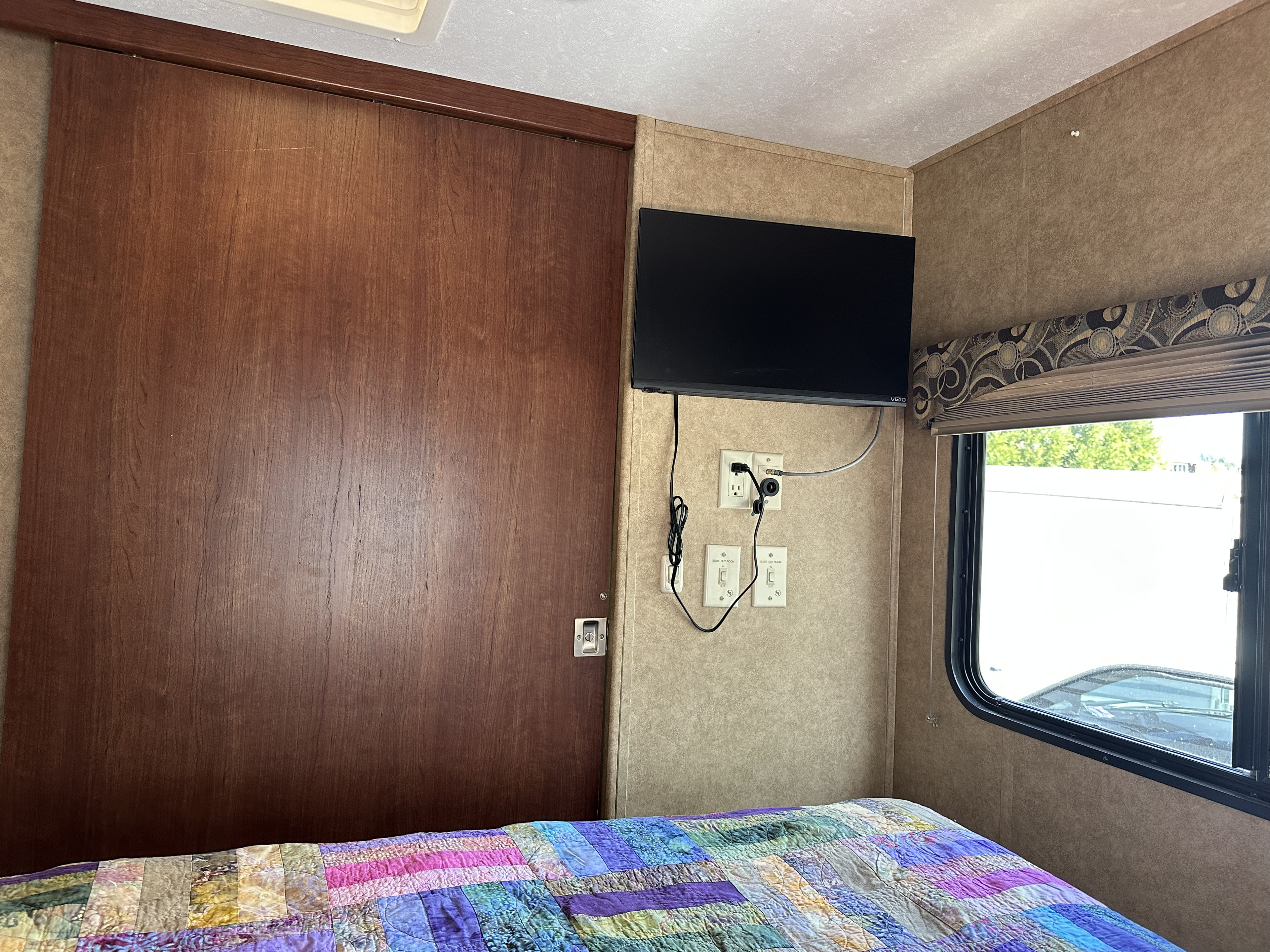 RV rental bedroom with mounted TV, slide-out controls, a wooden door, and a colorful quilted bed beside a window.. Forest River Sunseeker 2014