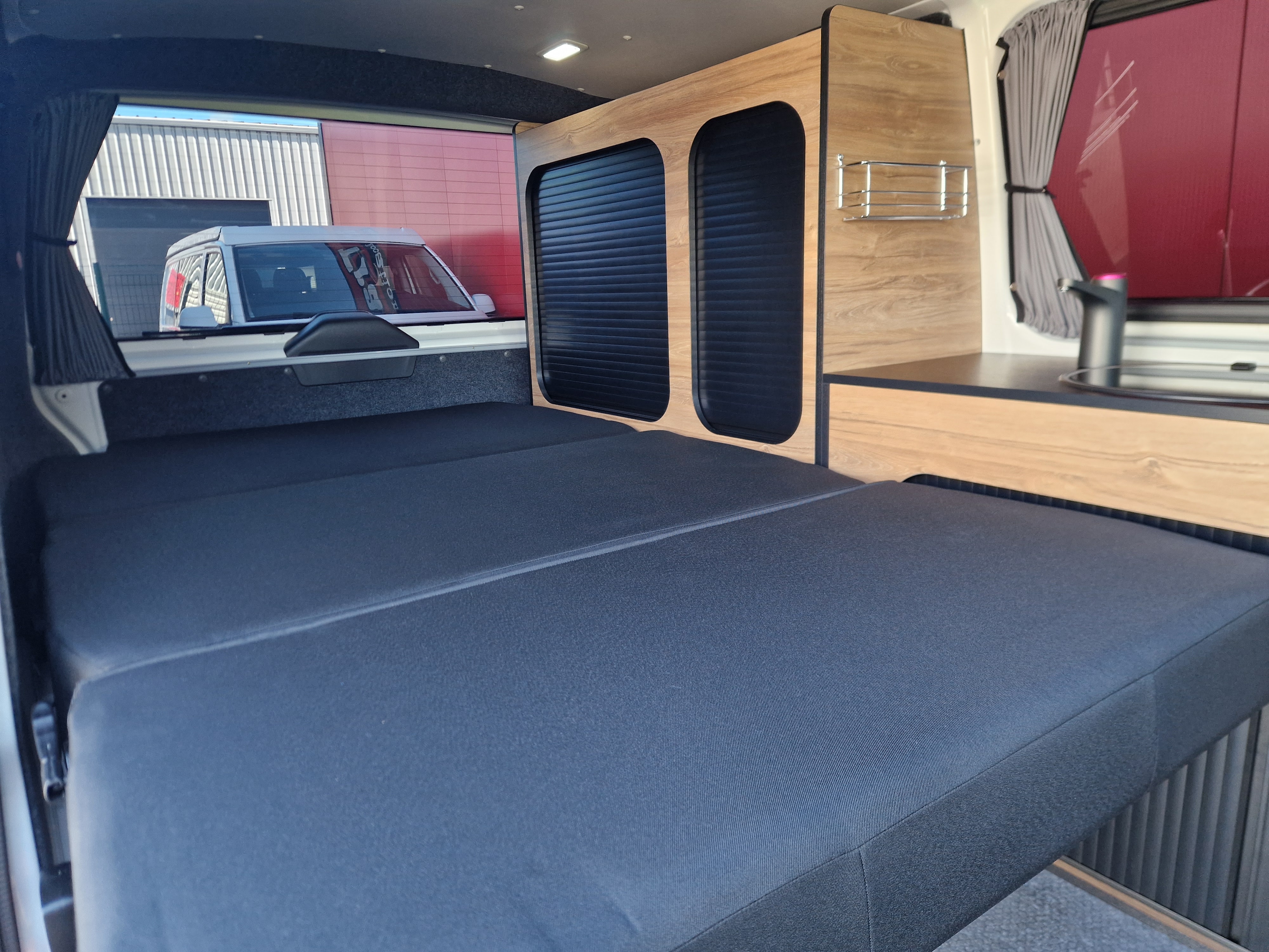 Cozy RV rental interior featuring a dark gray modular bed, wood cabinets, and a functional kitchenette.. Volkswagen Transporter T6 2025