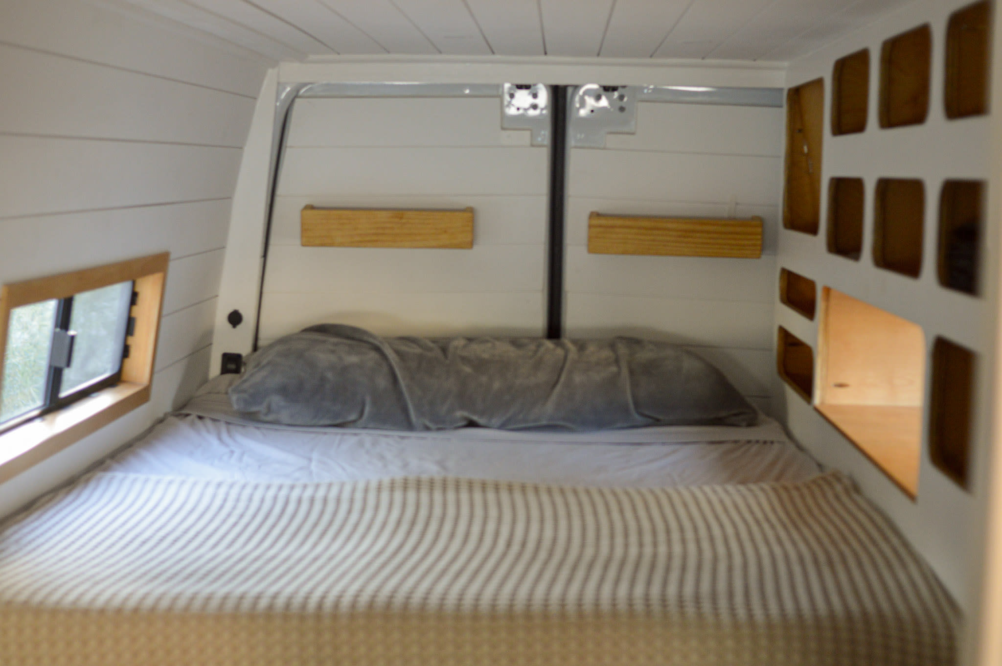 This RV rental shows a cozy bedroom setup with a striped bed, wooden shelves, and cubby storage built into white plank walls.. Mercedes-Benz Sprinter 2020