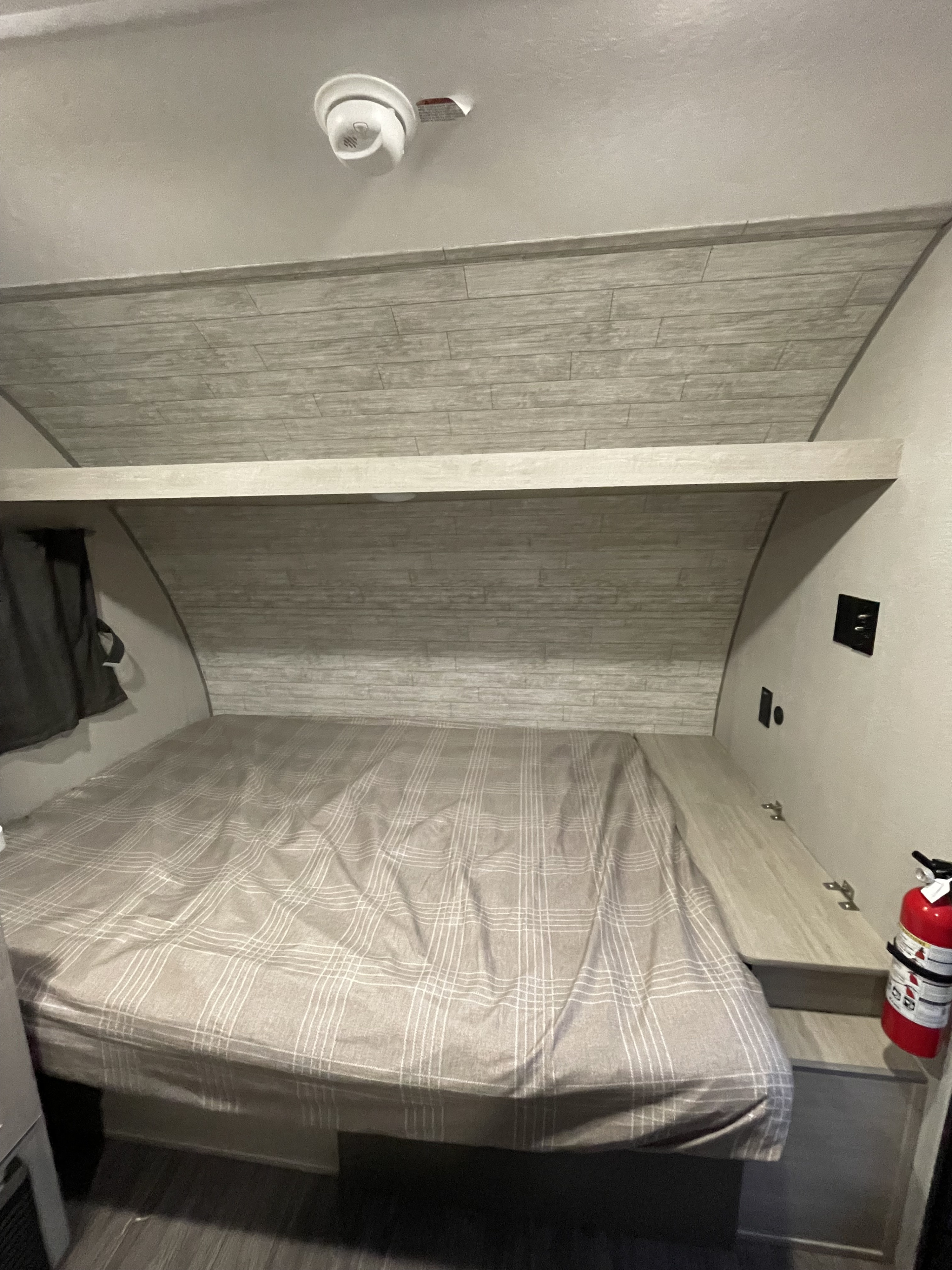 RV rental interior: bed with plaid cover, overhead shelf, smoke detector, and fire extinguisher.. Forest River Aurora ROK 2026