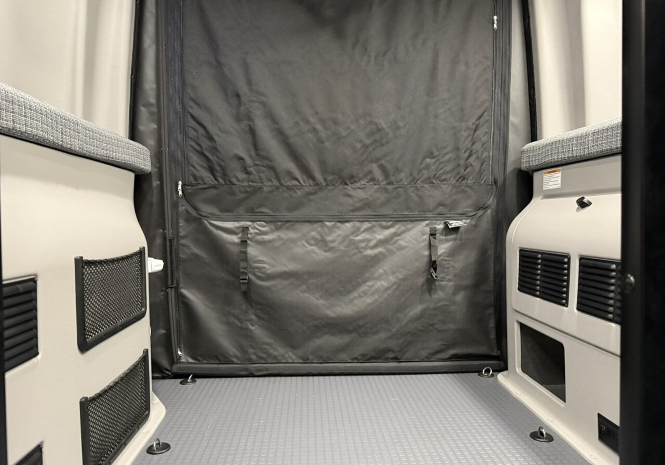 RV rental interior: Rear cargo space with a black fabric partition, bench seating, storage nets, and textured floor.. Winnebago Revel 2024